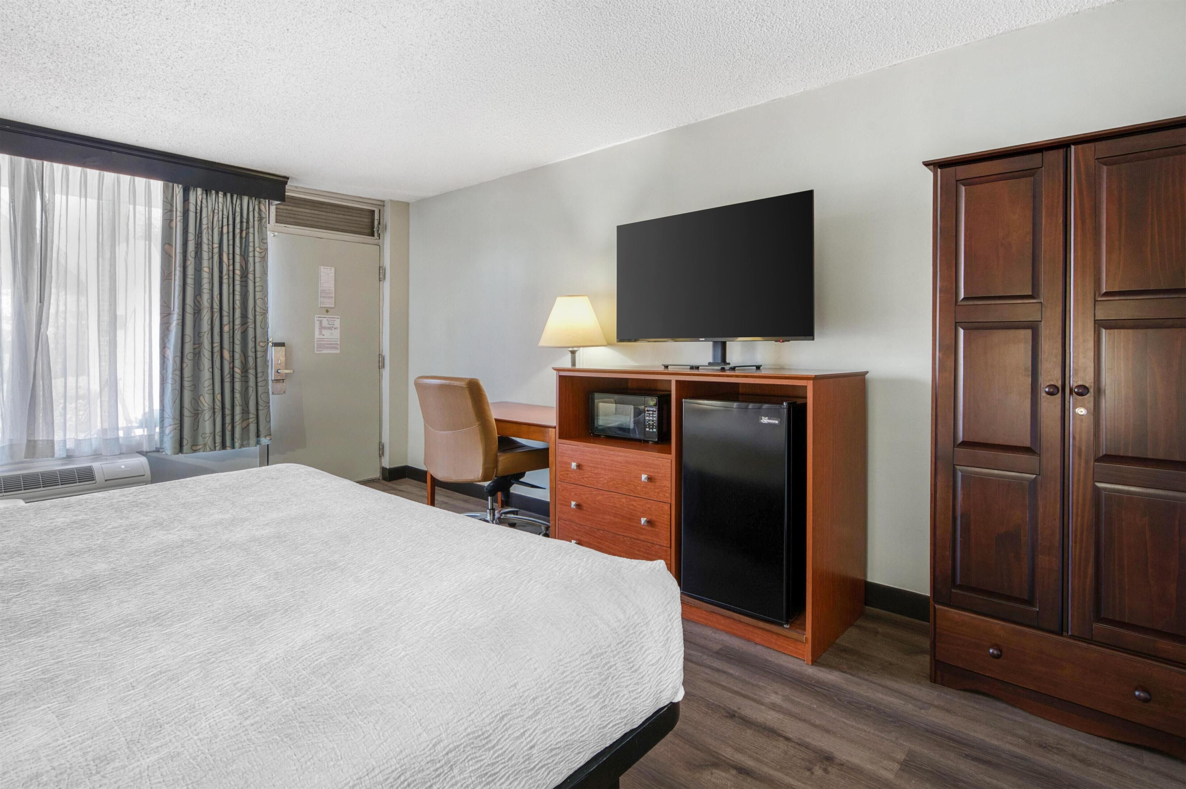 A hotel room with a bed, a desk with a television, a refrigerator, a chair, and a wardrobe.