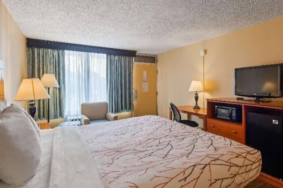 A hotel room with a bed, a desk with a television, a chair, and a desk lamp.