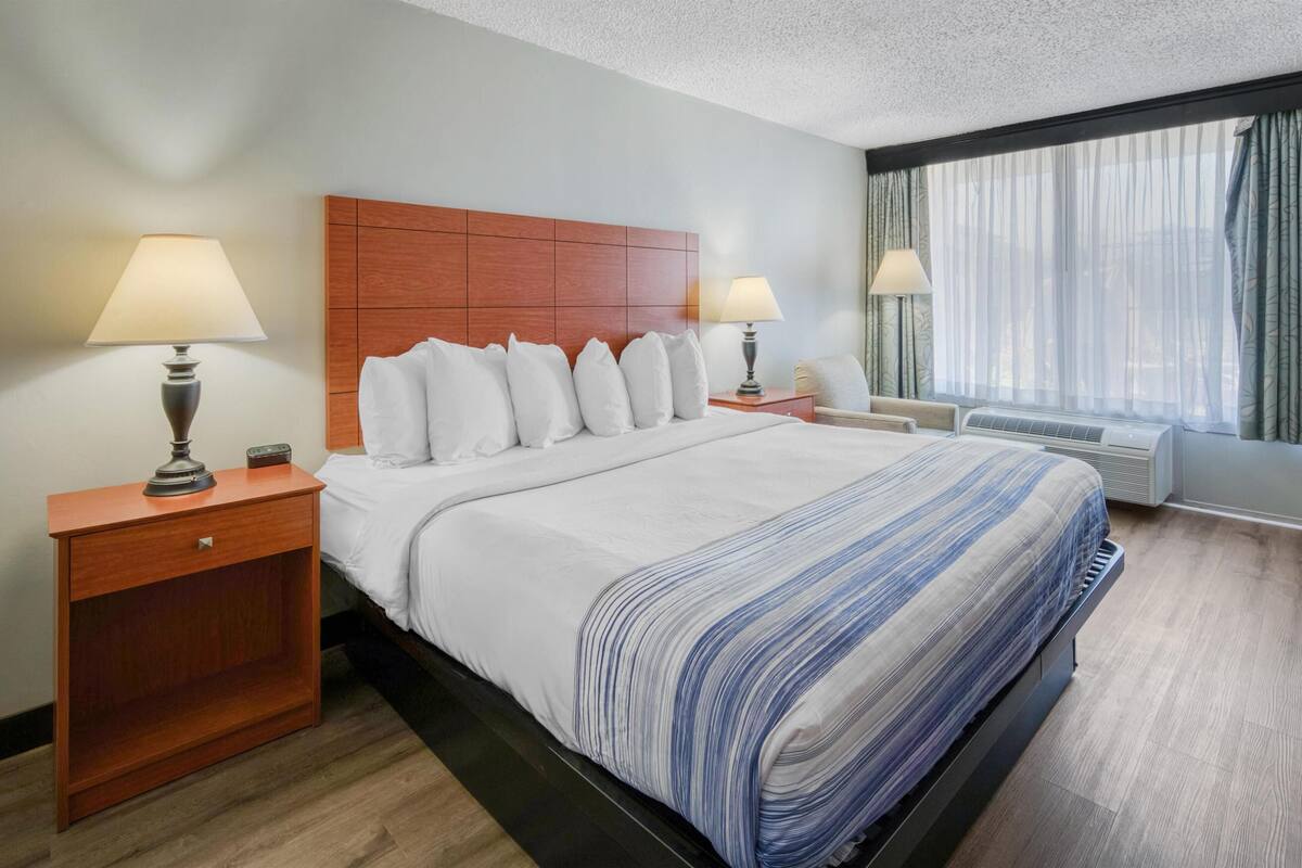 A hotel room with a large bed, two bedside tables with lamps, a wooden headboard, a window with curtains, and an air conditioning unit.