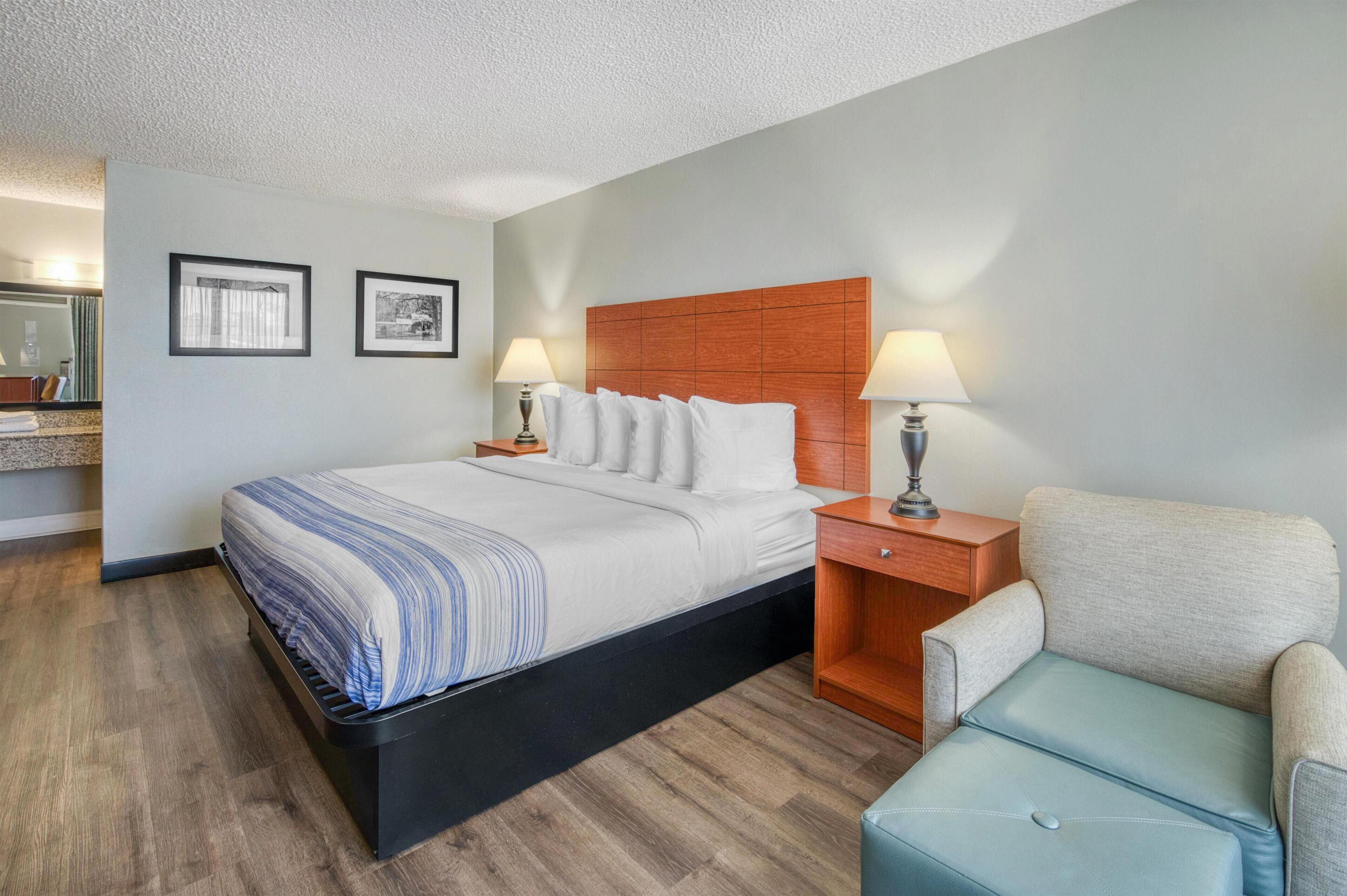 Una habitación de hotel con una cama, dos mesitas de noche con lámparas, una silla y cuadros en la pared.