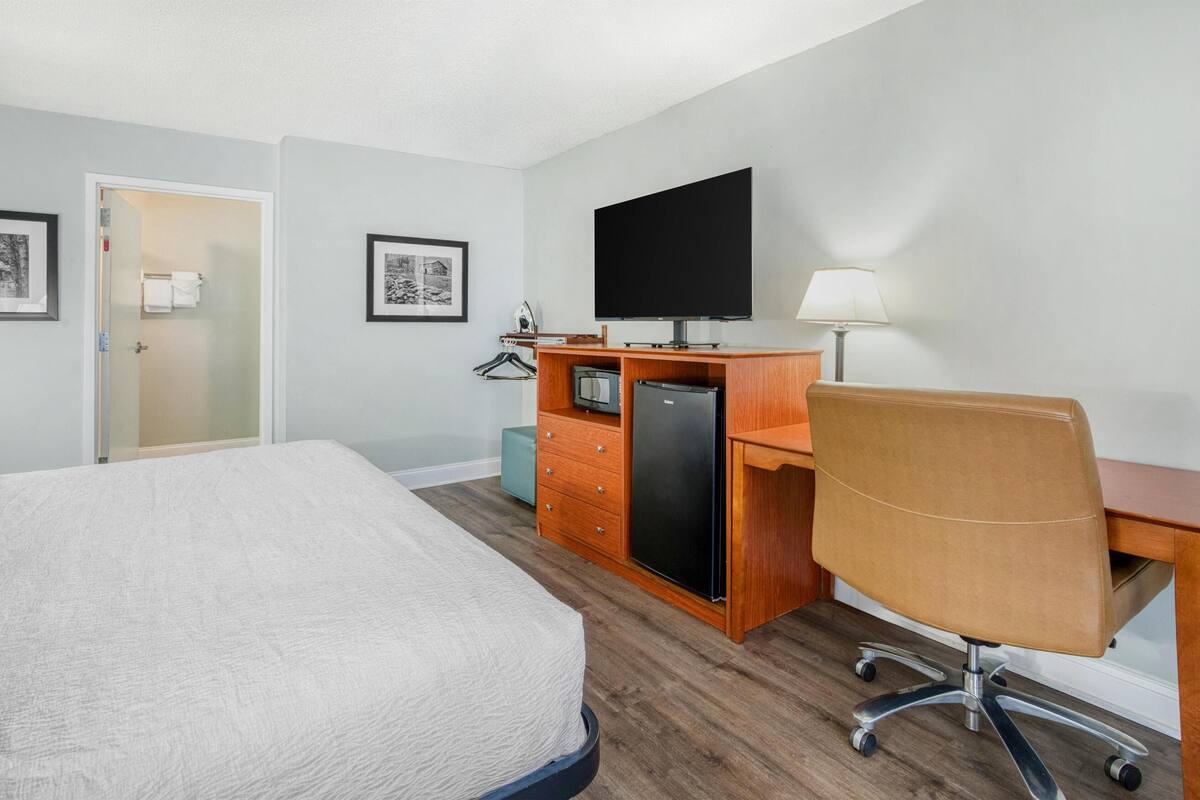 A hotel room with a bed, a desk with a computer, a flat-screen TV, a lamp, and a refrigerator.