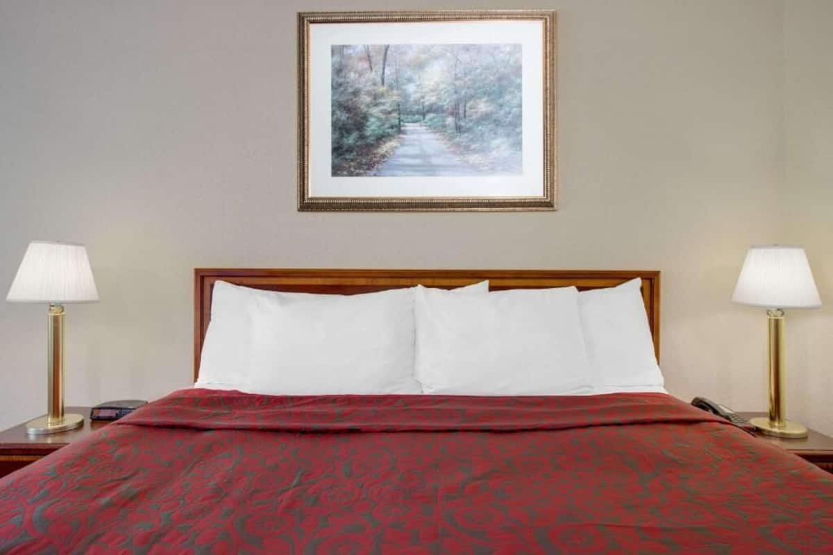 A neatly made bed with a red patterned bedspread, two bedside lamps, and a framed picture of a forest path on the wall.