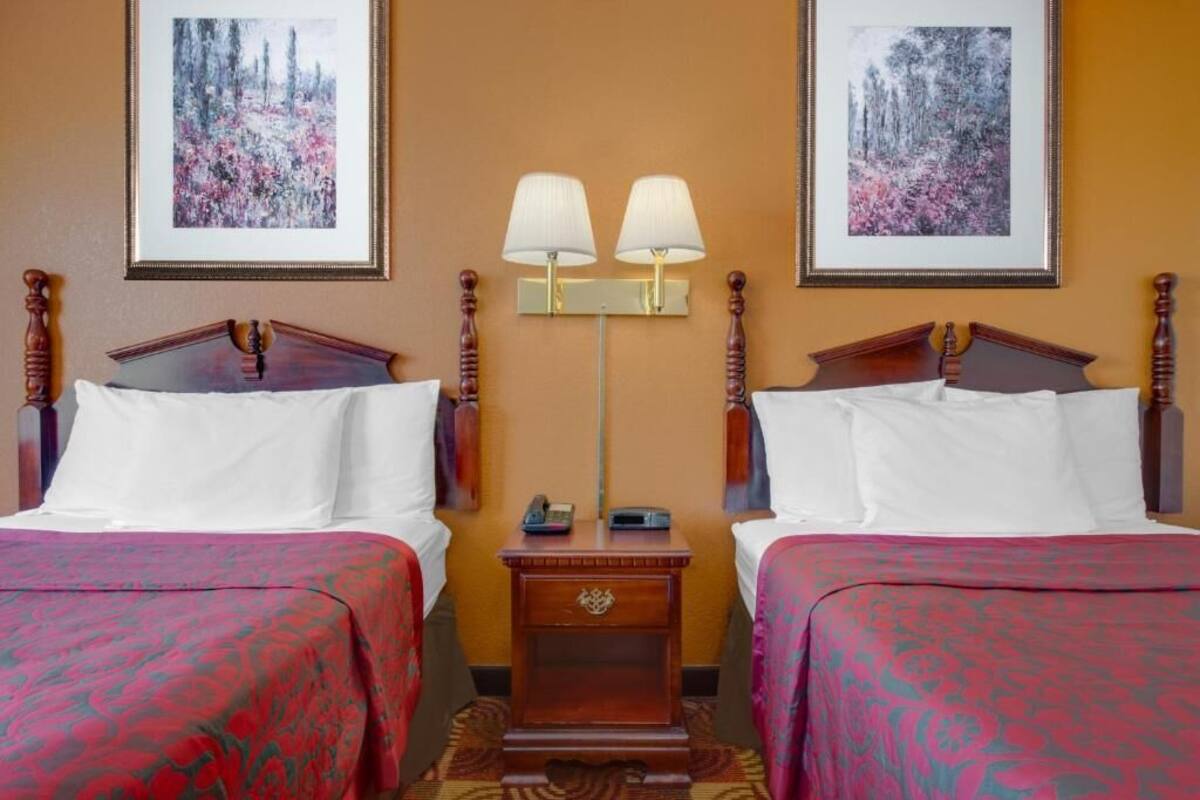 A hotel room with two beds, each with a wooden headboard, white bedding, and a red patterned bedspread. There is a nightstand between the beds with a lamp and a phone. Two framed pictures hang on the wall above the beds.