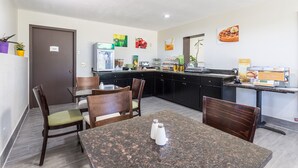 A modern kitchen with granite countertops, wooden chairs, and a dining table. There are various food items and a sign indicating hot beverages.