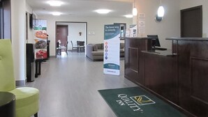 A hotel reception area with a green carpet, a reception desk, a seating area with chairs, and a poster advertising the hotel.