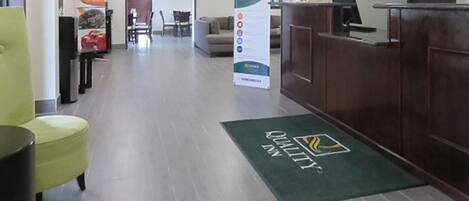 Zona de recepción de un hotel con alfombra verde, un mostrador de recepción, un área de estar con sillas y un cartel publicitario del hotel.