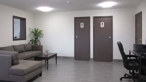 A modern waiting area with a grey sofa, a small table, and a black office chair. There are two doors with signs indicating restrooms.