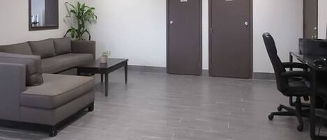 Un área de espera moderna con un sofá gris, una mesita y una silla de oficina negra. Hay dos puertas con indicaciones de baños.