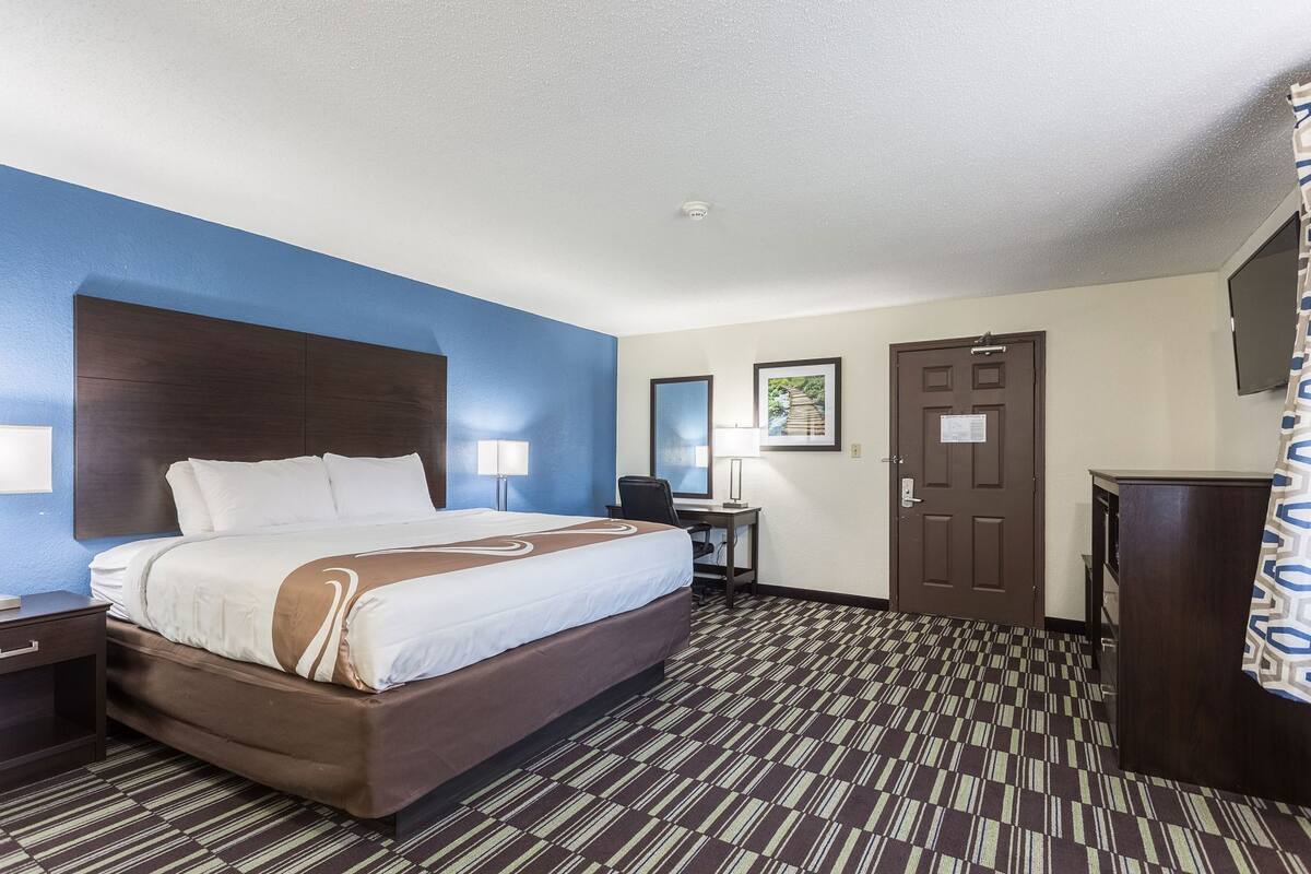 A hotel room with a large bed, a desk, a chair, a TV, and a door.