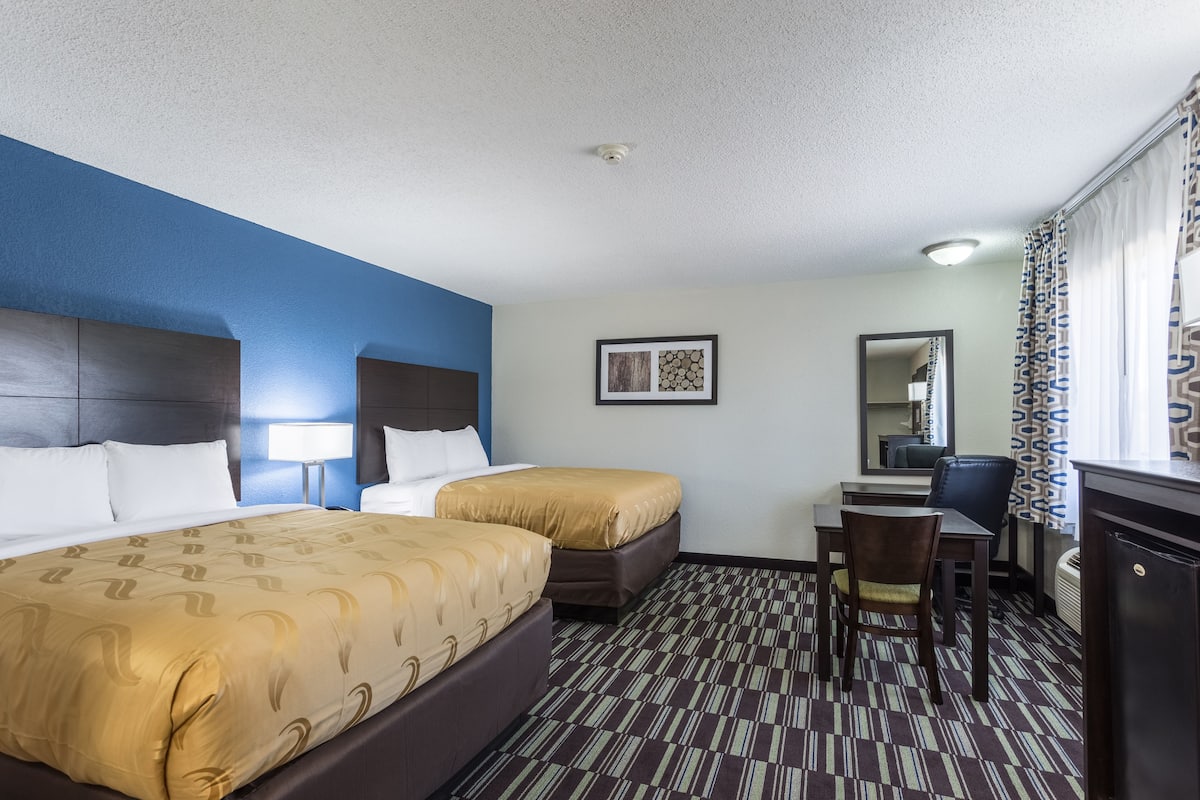 A hotel room with two beds, a desk, a chair, and a small kitchenette.