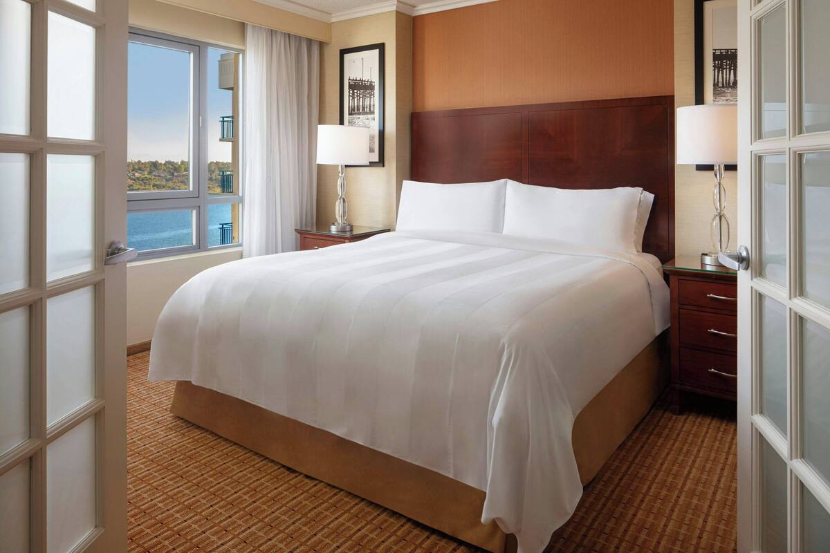 A hotel room with a large bed, wooden headboard, and a view of the sea through a large window.