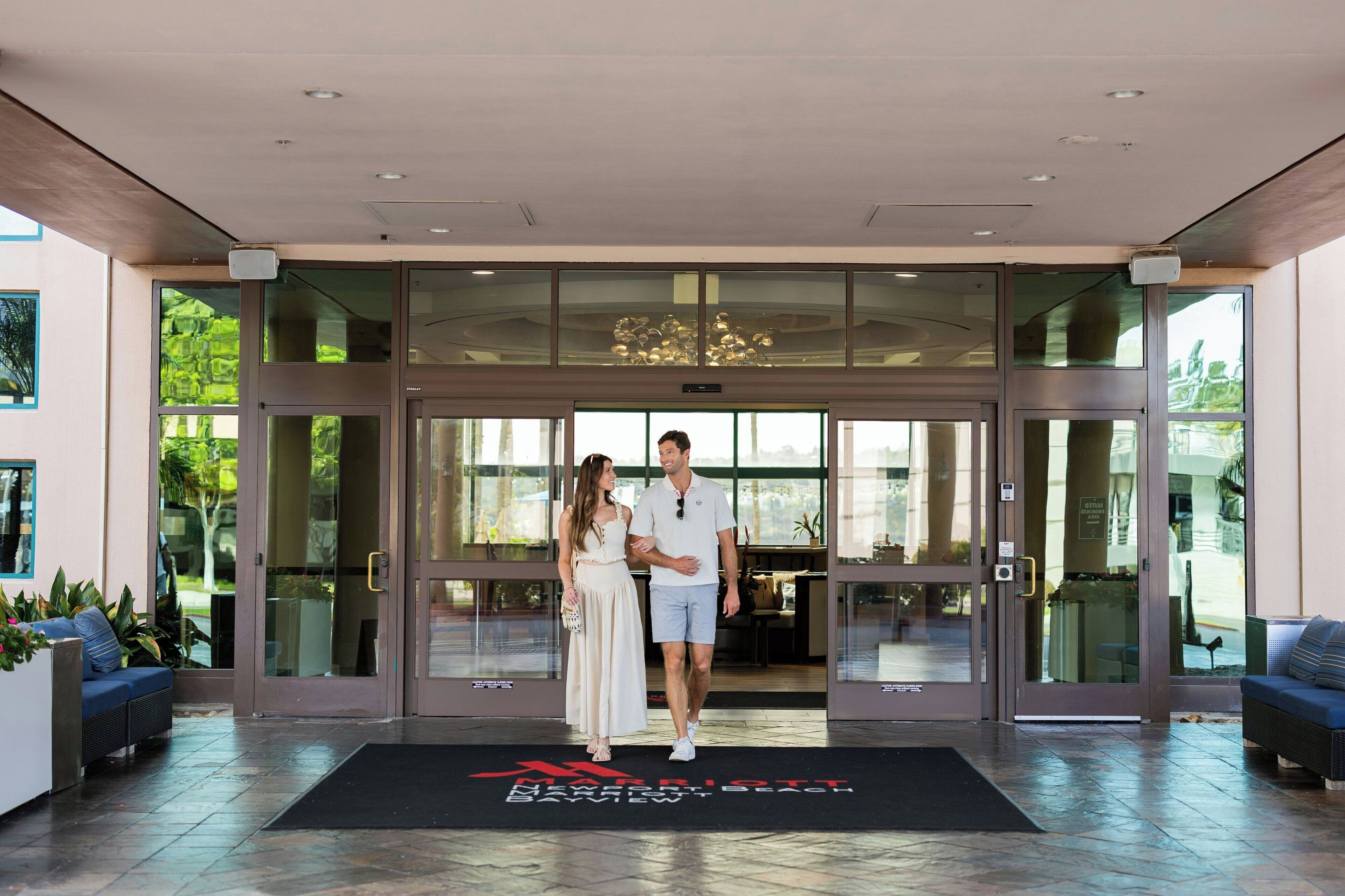 A hotel entrance with glass doors, a black mat with red text, and outdoor seating.