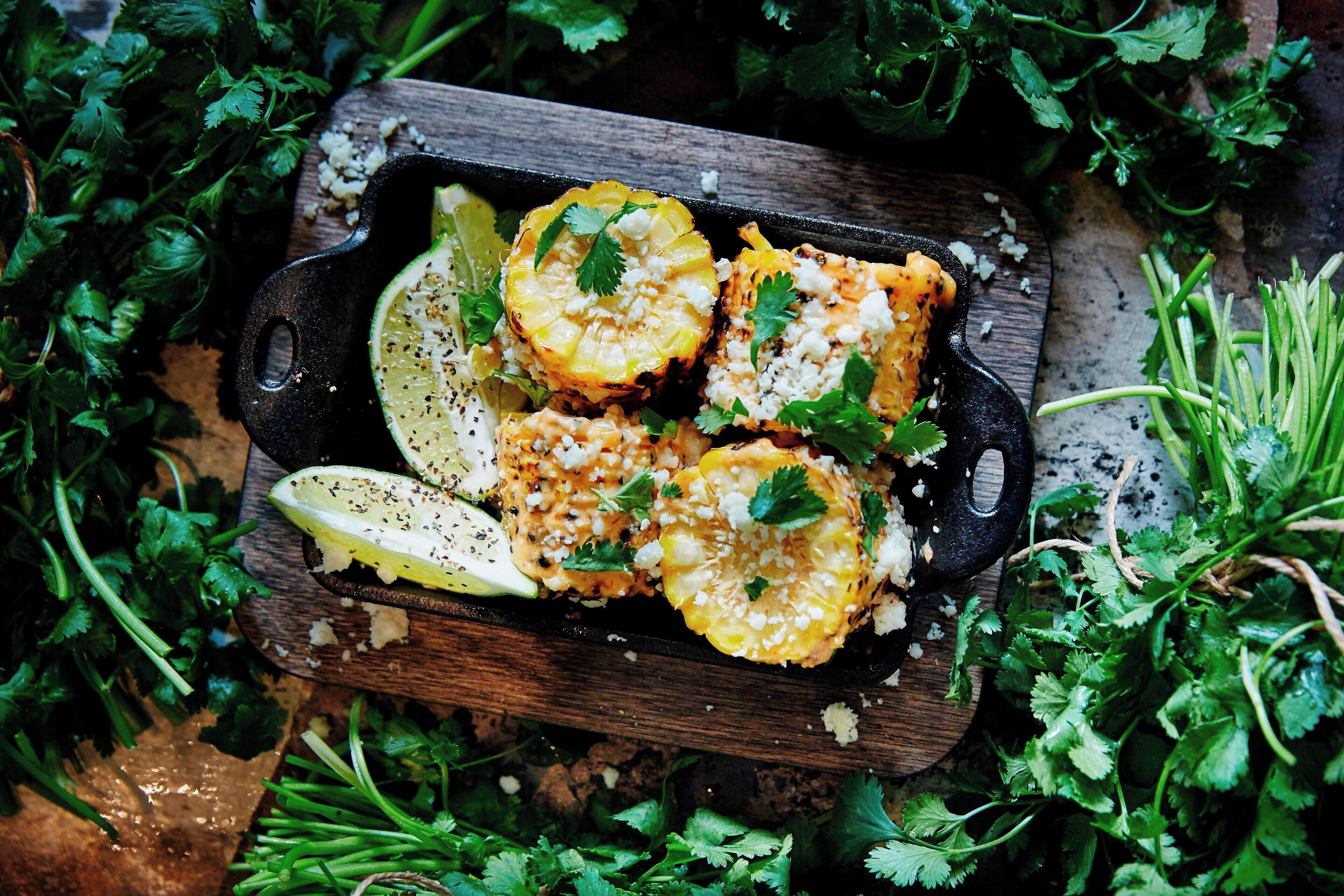 A cast iron skillet with grilled corn on the cob, garnished with cilantro and lime, surrounded by fresh herbs.