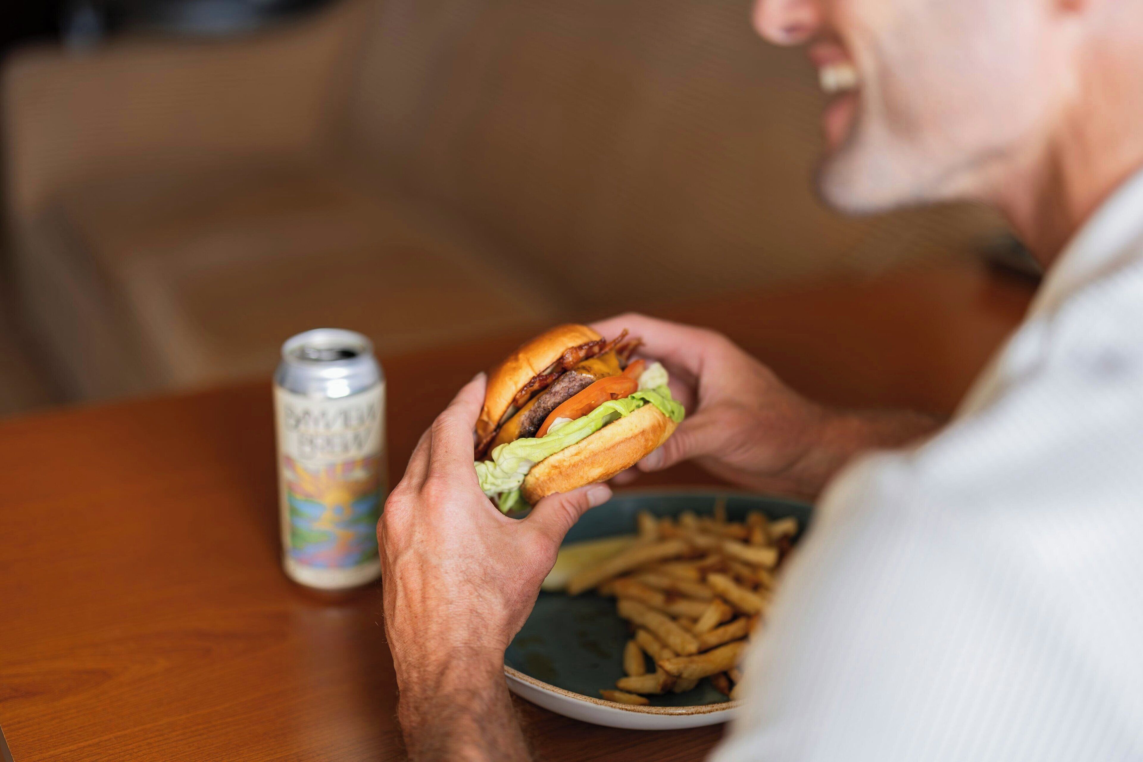 A person holding a burger and a can of beer on a table with a plate of fries.
