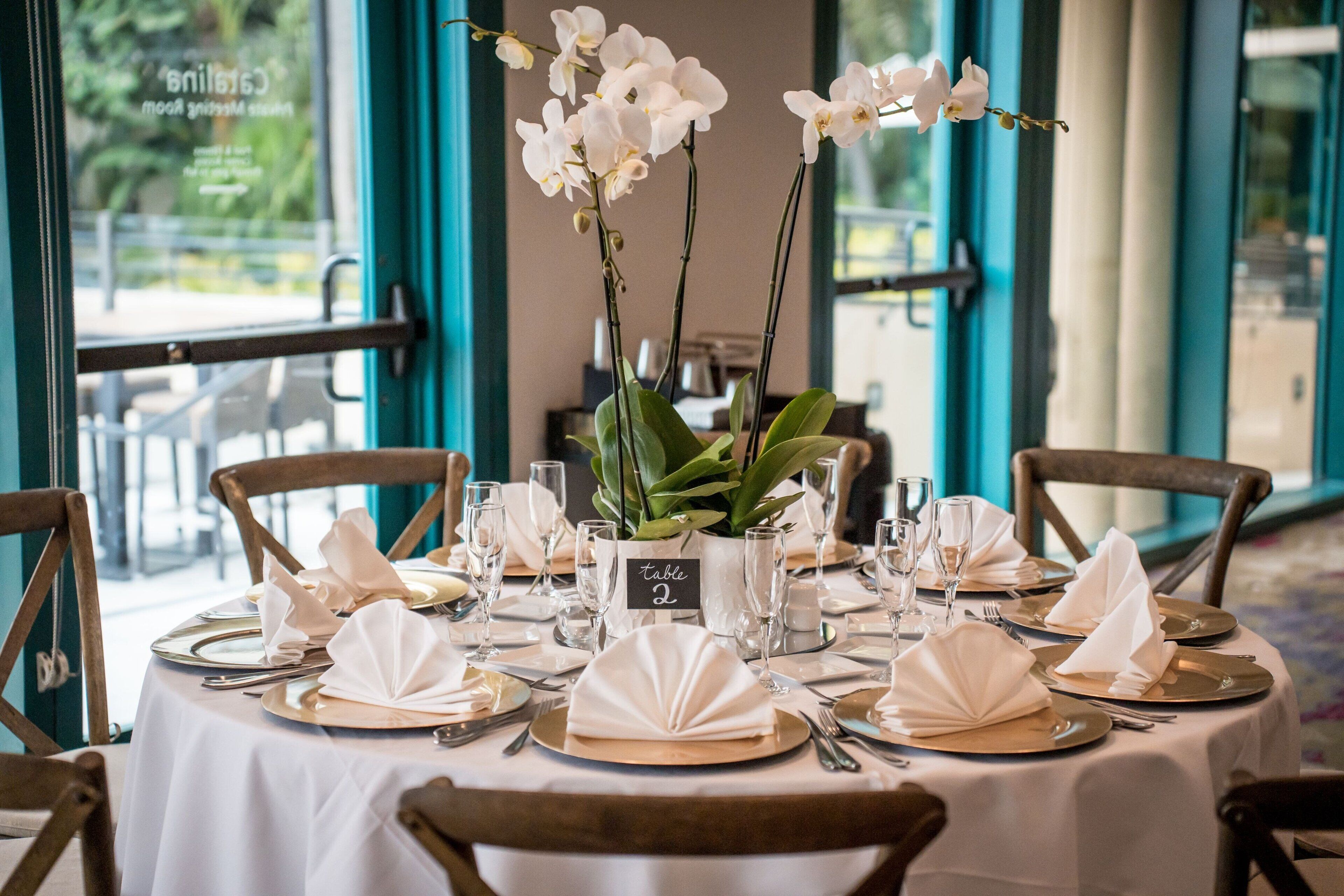 A dining table set for four with white tablecloths, folded napkins, and a vase of white orchids.