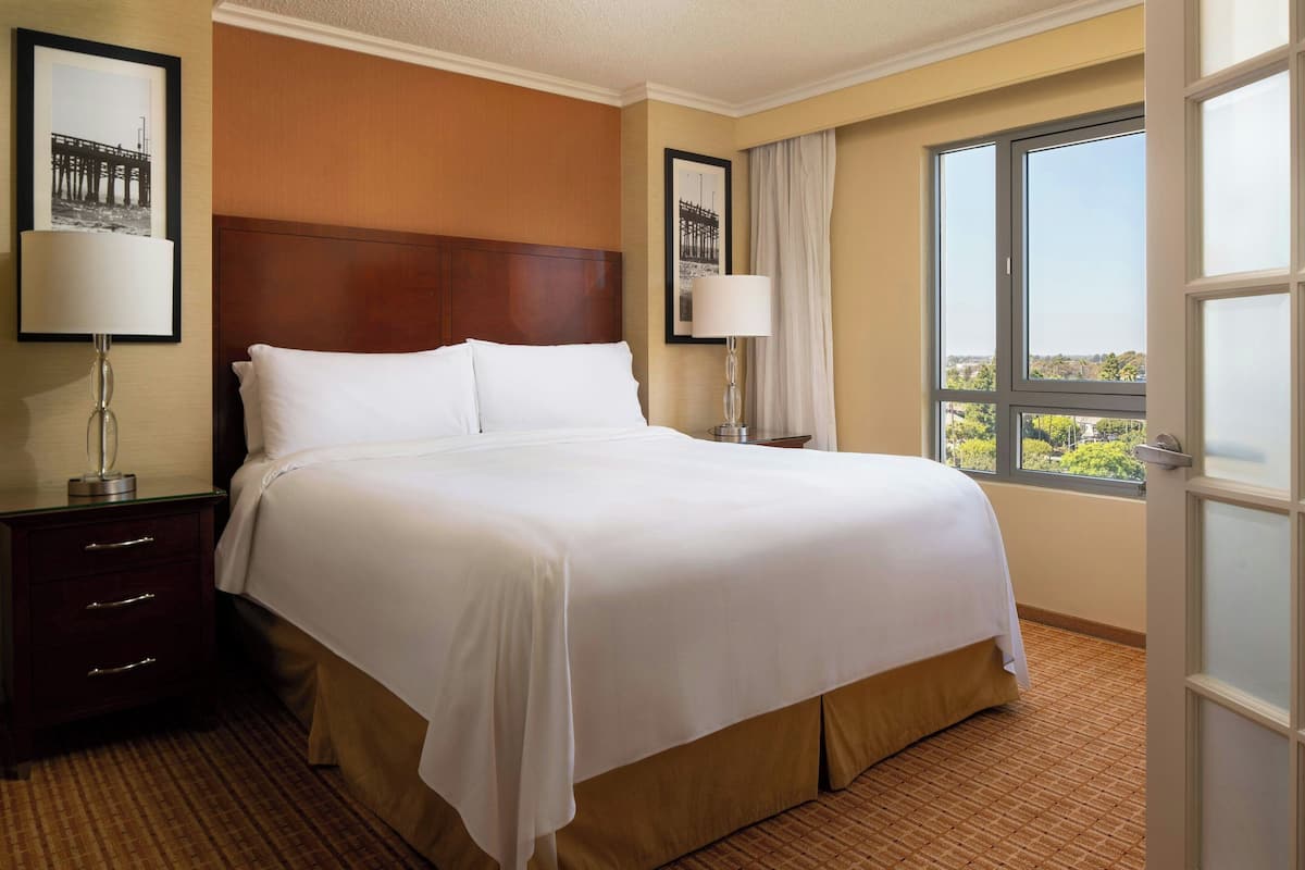 A hotel room with a large bed, two bedside tables with lamps, a view of a cityscape through a large window, and a framed picture on the wall.