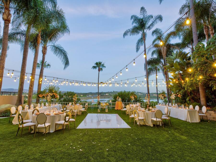 Un lugar para bodas al aire libre con pista de baile, mesas, sillas y luces colgantes.