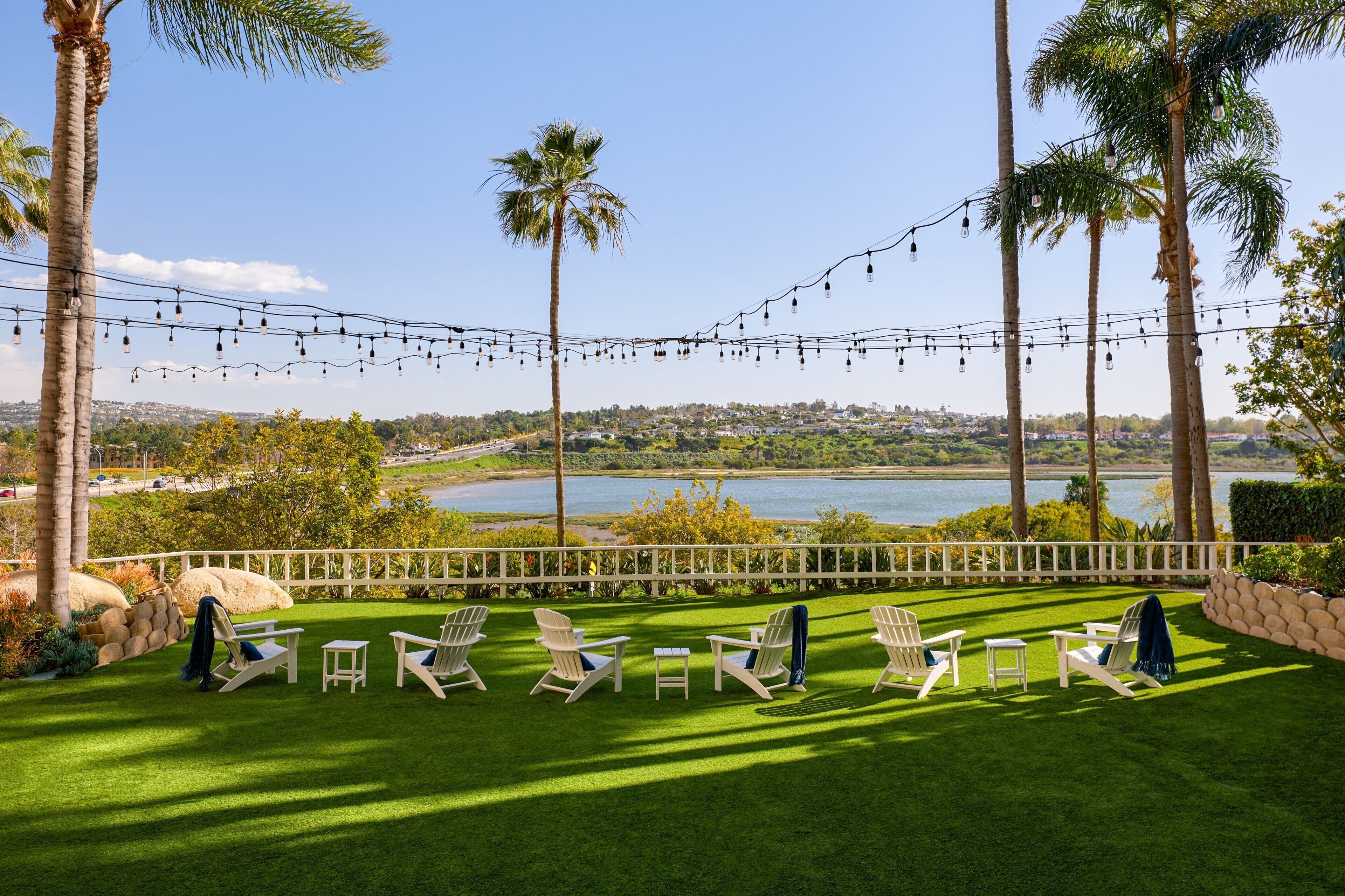 A patio with lawn chairs and a table, overlooking a lake with palm trees and a white fence.