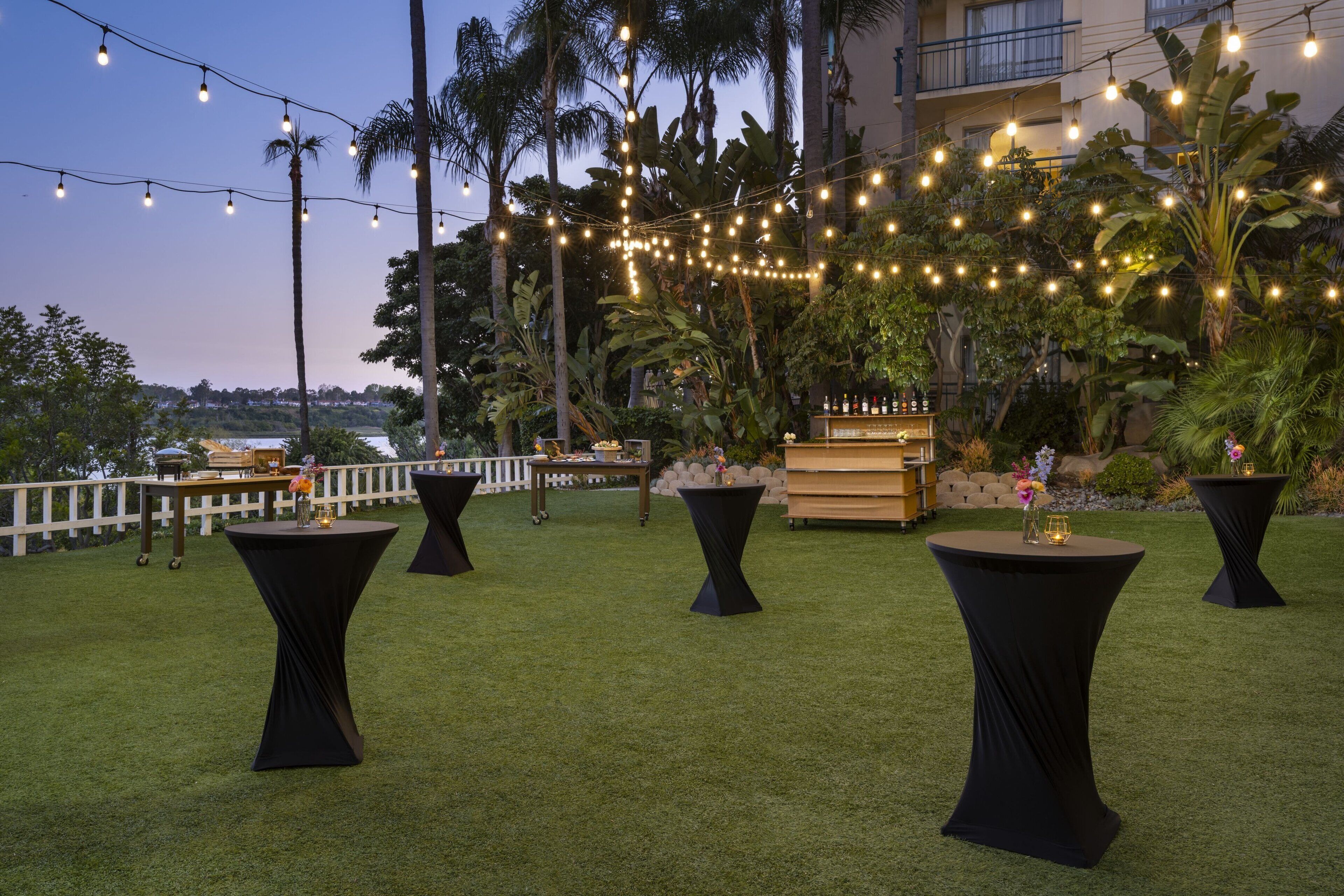 An outdoor seating area with round tables and chairs, string lights, and a view of a building and palm trees.