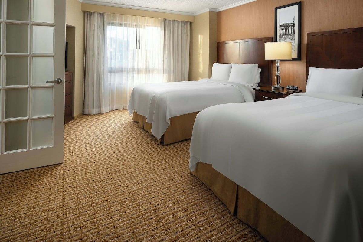 A hotel room with two beds, a wooden headboard, a bedside table with a lamp, and a large window with sheer curtains.
