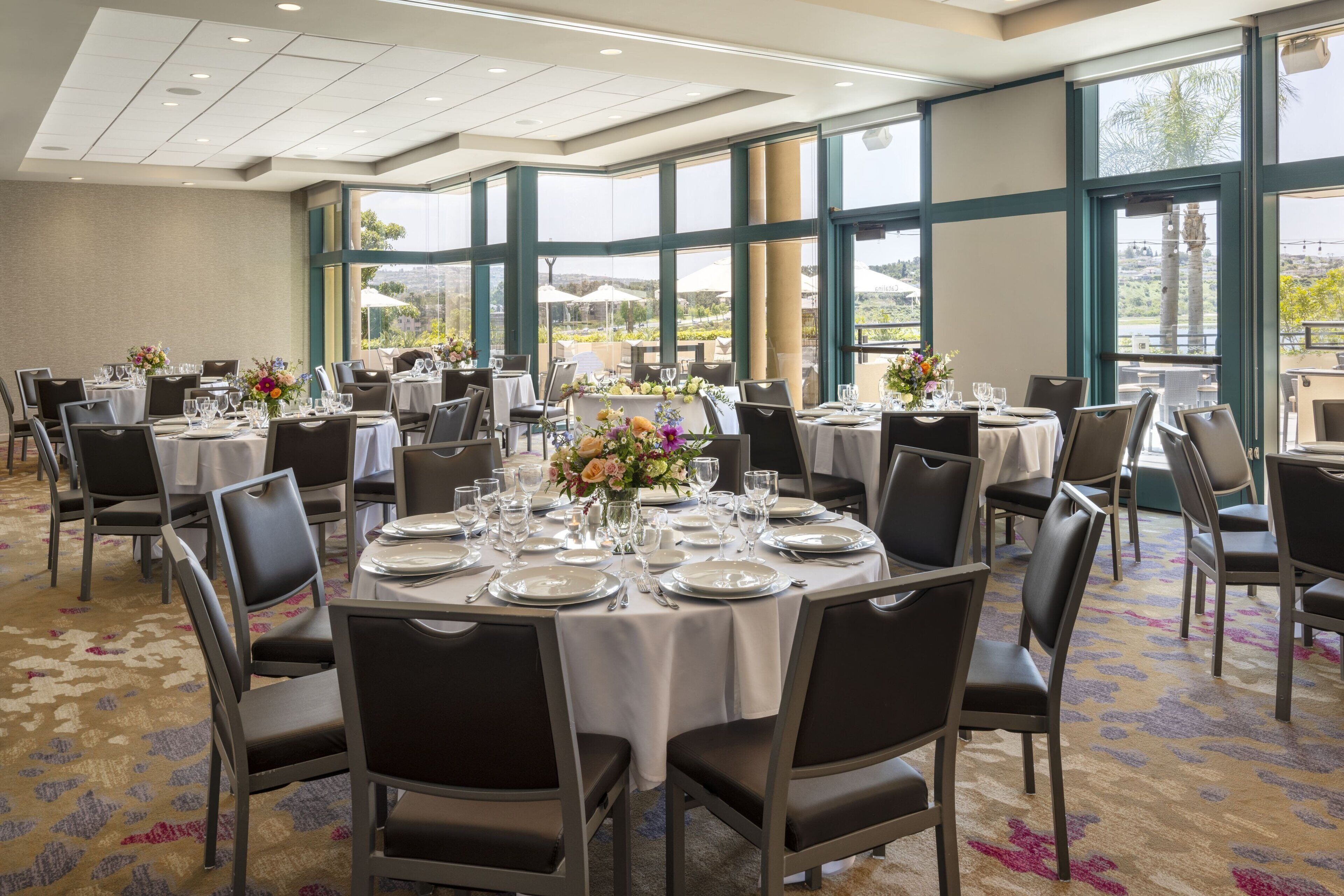 Un salón de banquetes con mesas redondas dispuestas para comer, ventanas amplias con vistas y suelo alfombrado.