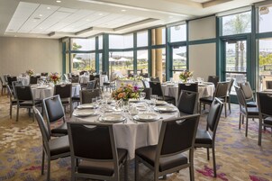Un salón de banquetes con mesas redondas dispuestas para comer, ventanas amplias con vistas y suelo alfombrado.
