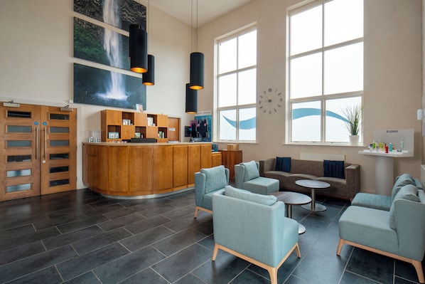 A modern hotel lobby with a reception desk, seating area, and large windows.