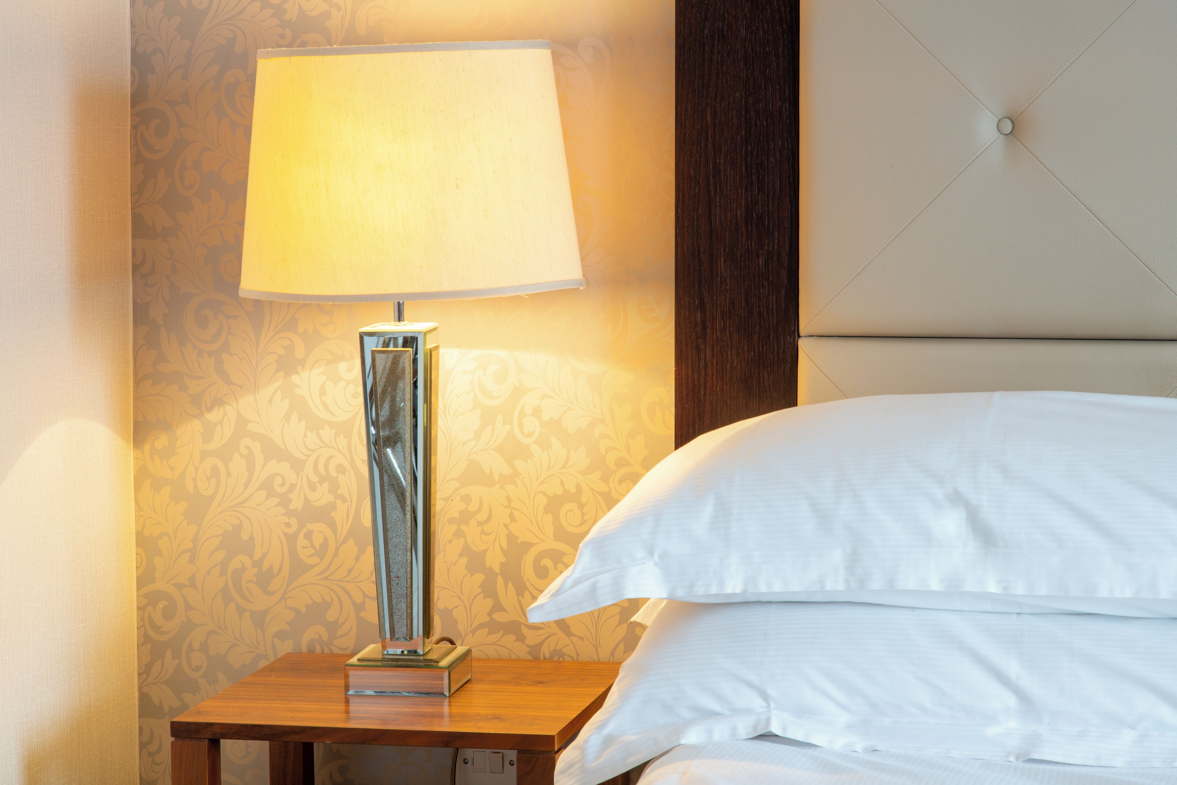 A neatly made bed with white linens, a bedside lamp on a wooden nightstand, and a patterned wallpaper in the background.