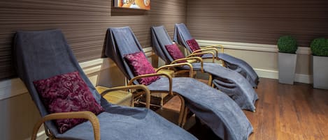 A row of wicker lounge chairs with blue covers and purple pillows, arranged in a spa-like setting with wooden flooring and a wall-mounted poster.