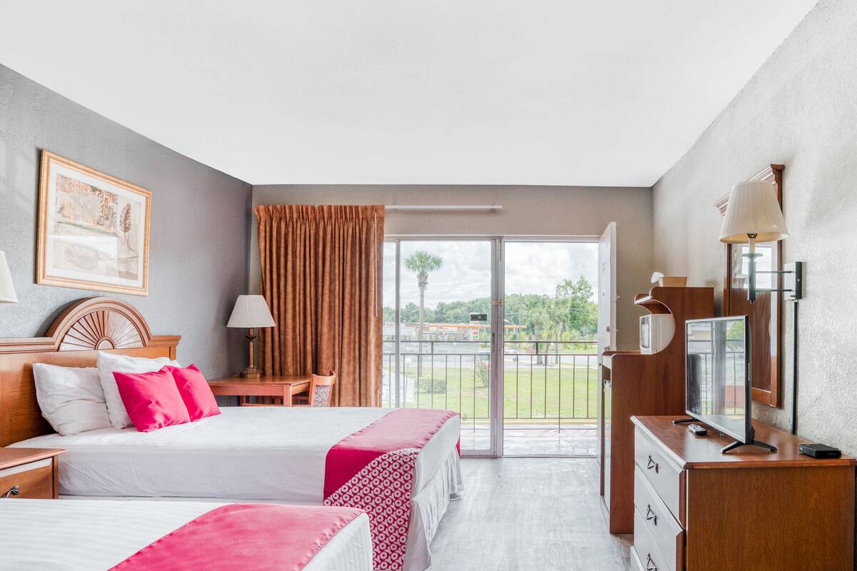 A hotel room with two beds, a desk, a TV, and a balcony with a view of greenery.