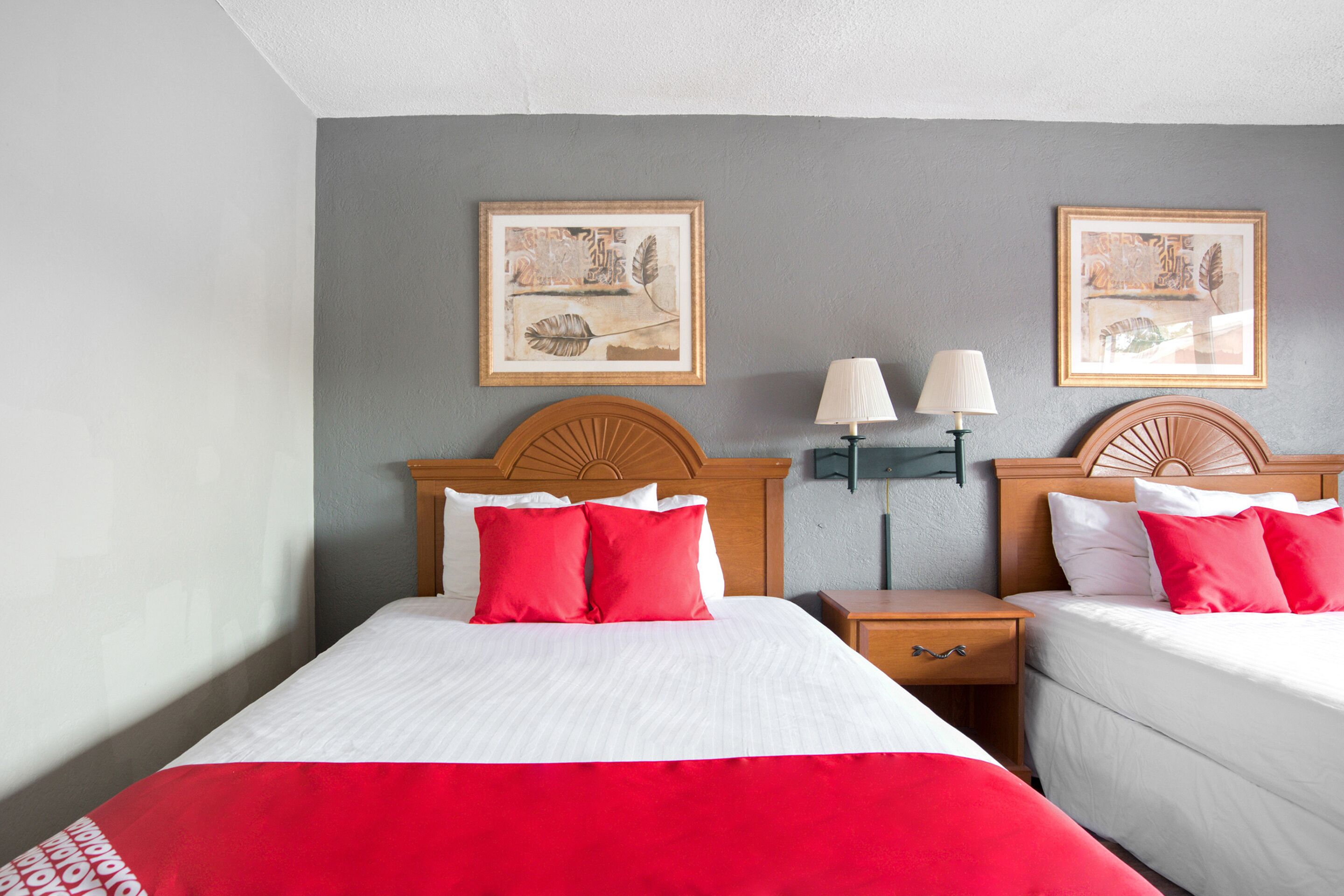 Quarto de hotel com duas camas, cada uma com cabeceira de madeira, roupa de cama branca e almofadas vermelhas. Há duas luminárias fixadas na parede e duas obras de arte emolduradas sobre as camas.