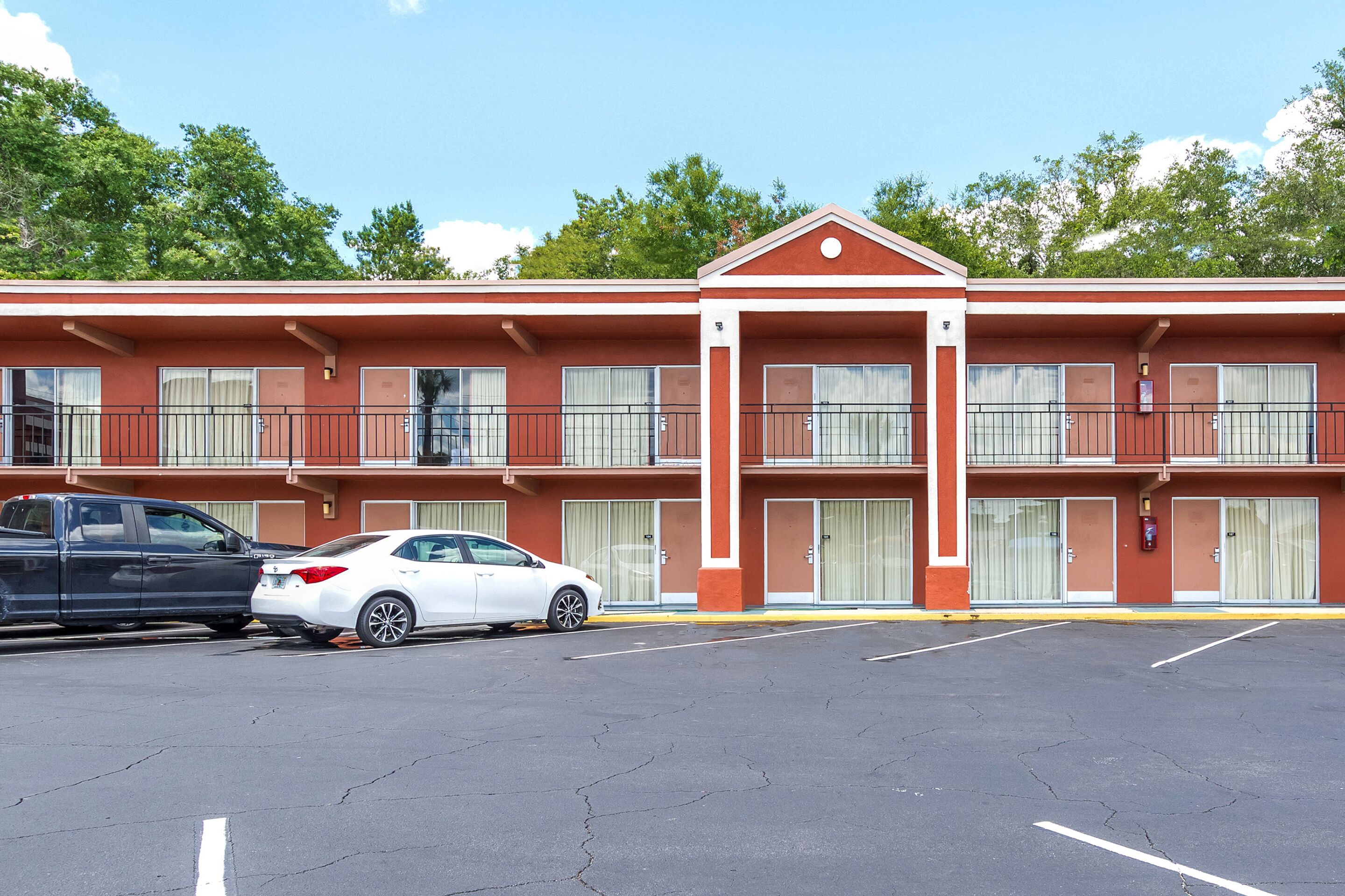 A motel with two-story architecture, featuring a central entrance with a red roof, multiple rooms with closed curtains, and a parking lot with two cars.