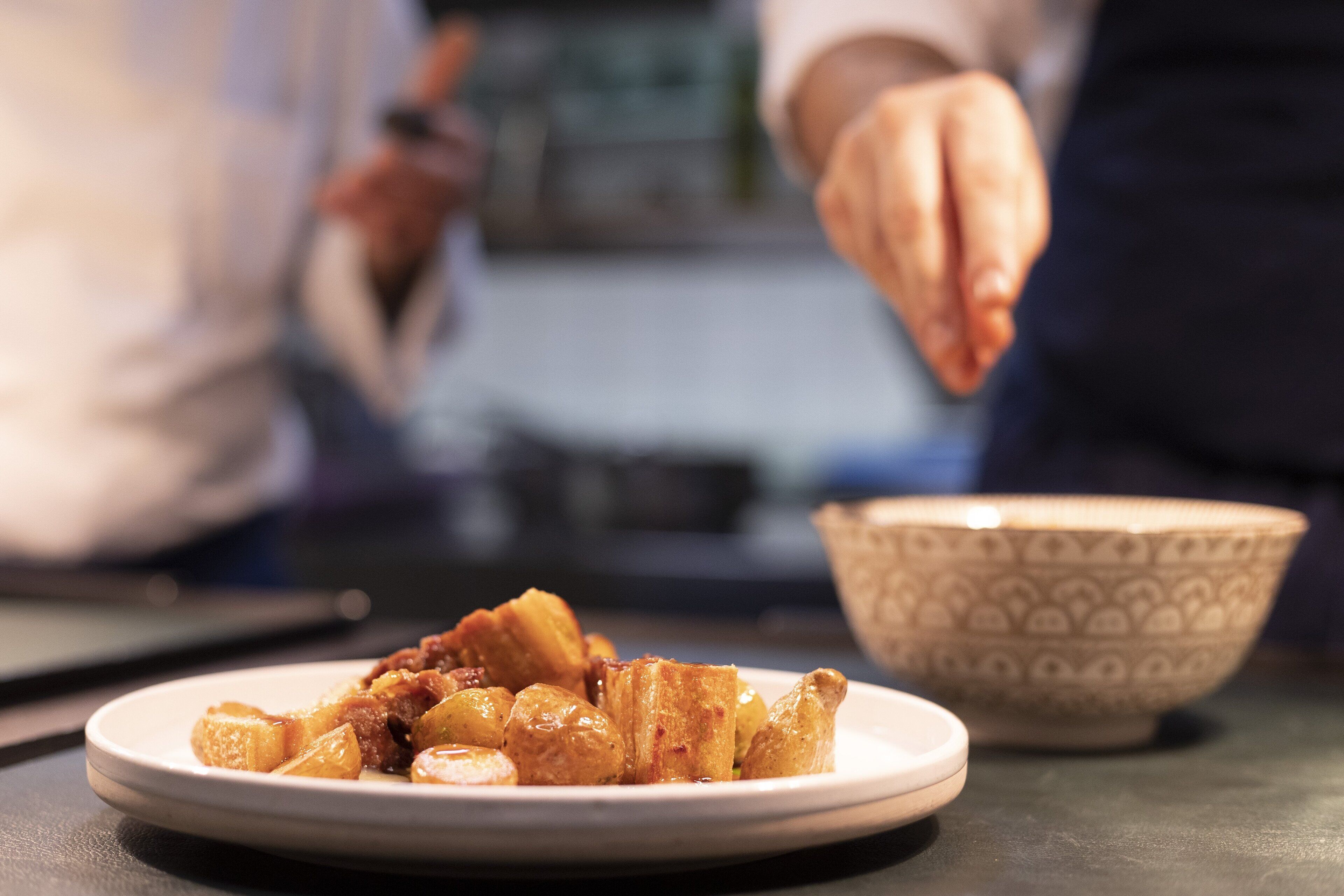 A plate of food with a hand reaching for it, a patterned bowl, and a blurred background.