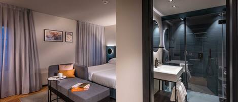 Una habitación de hotel moderna con sofá, una mesita con una taza y un libro, cama y zona de ducha con mampara y lavamanos.
