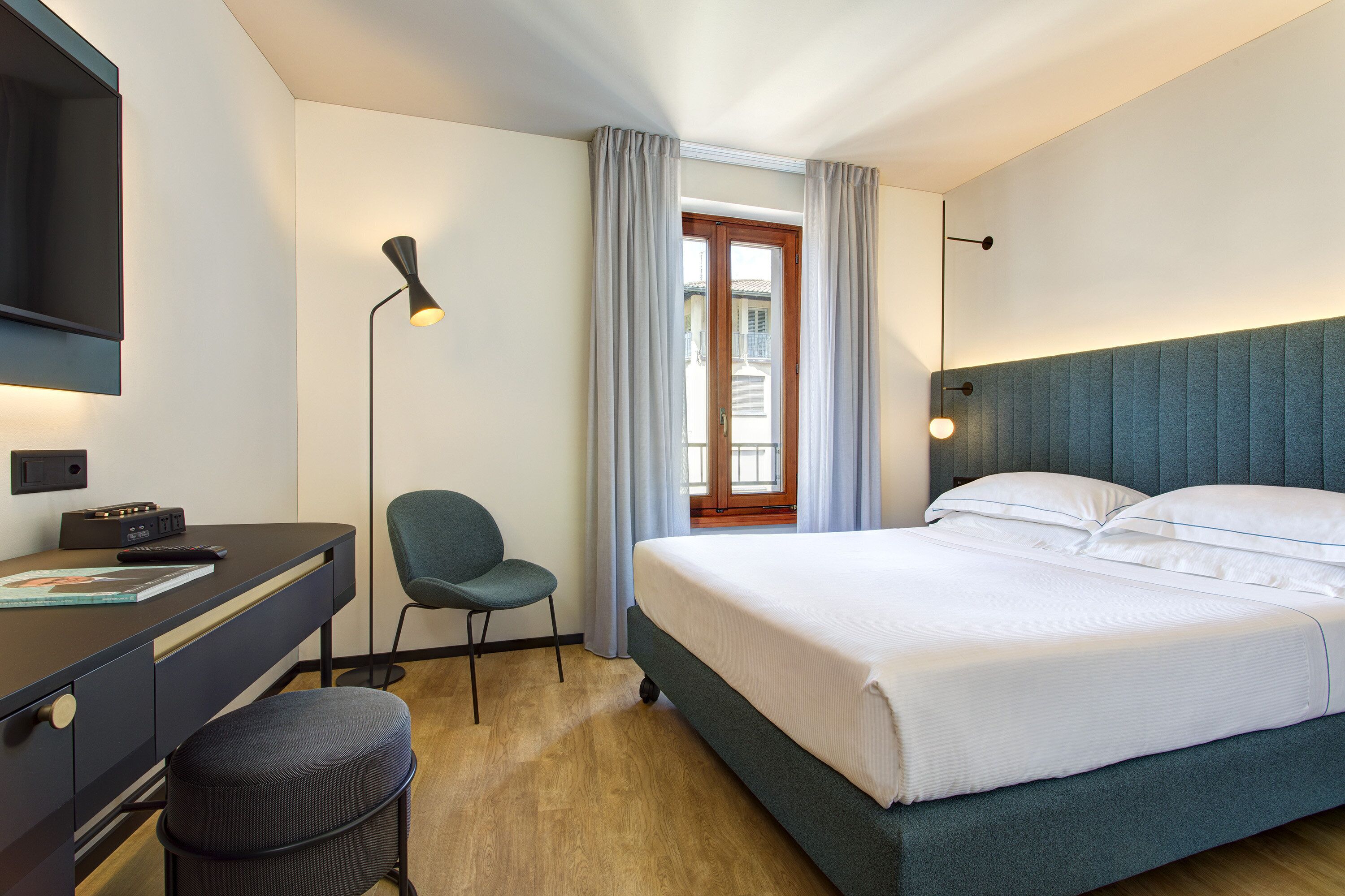 Habitación de hotel con cama, escritorio, silla, televisor y ventana con cortinas.