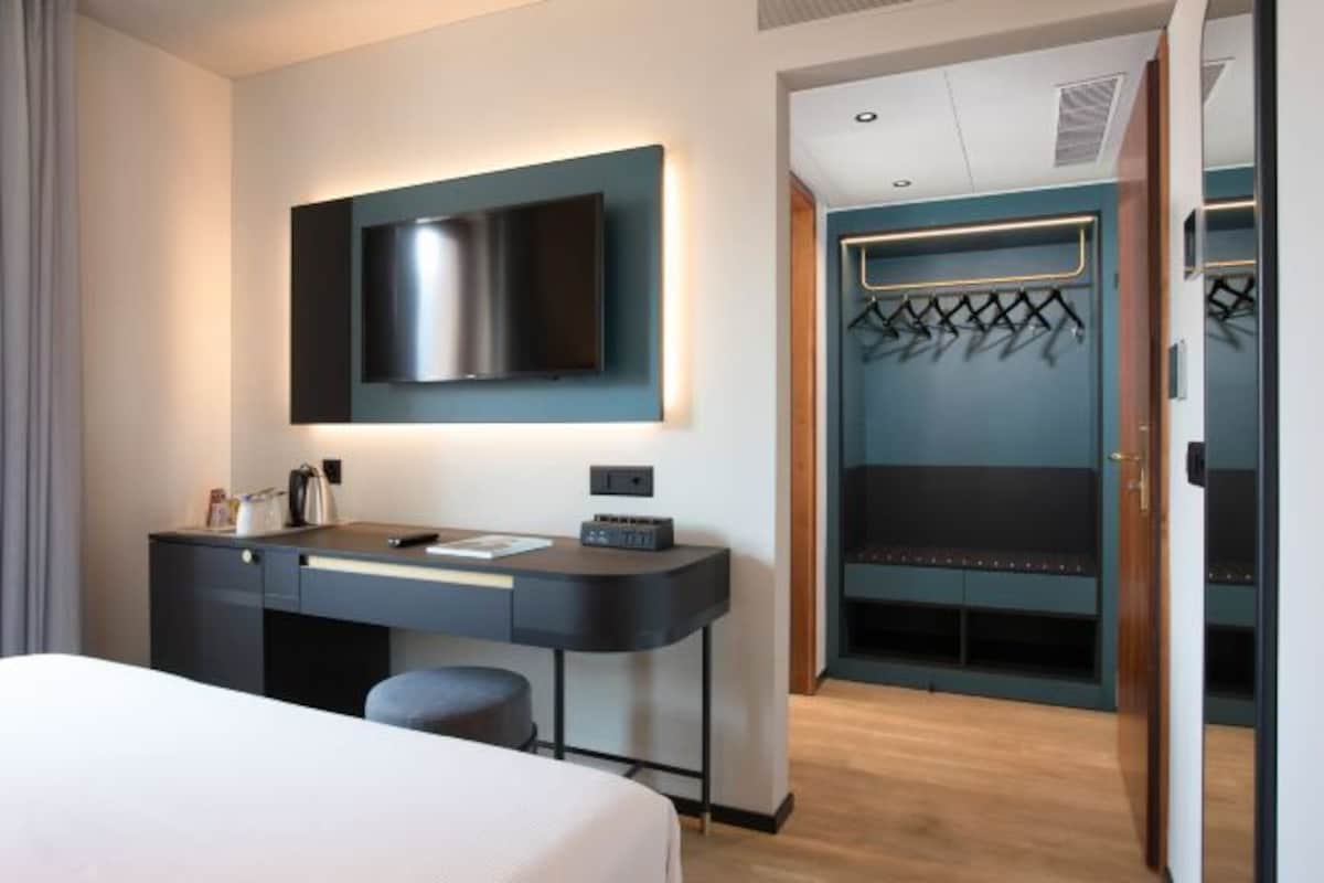 A hotel room with a bed, a desk with a flat-screen TV, a coat rack, and a wardrobe.