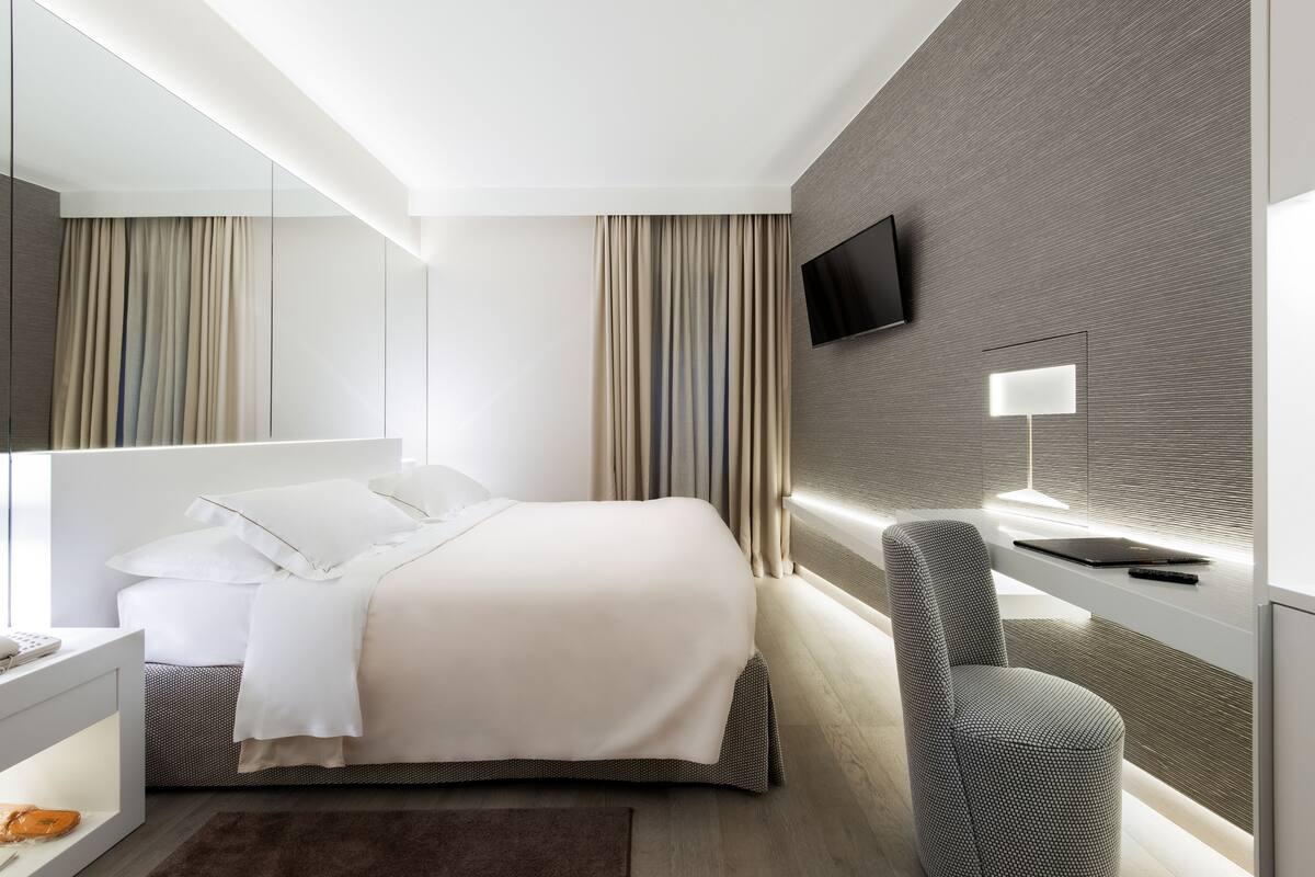 A modern hotel room with a large bed, a desk with a lamp, a TV mounted on the wall, and a chair.