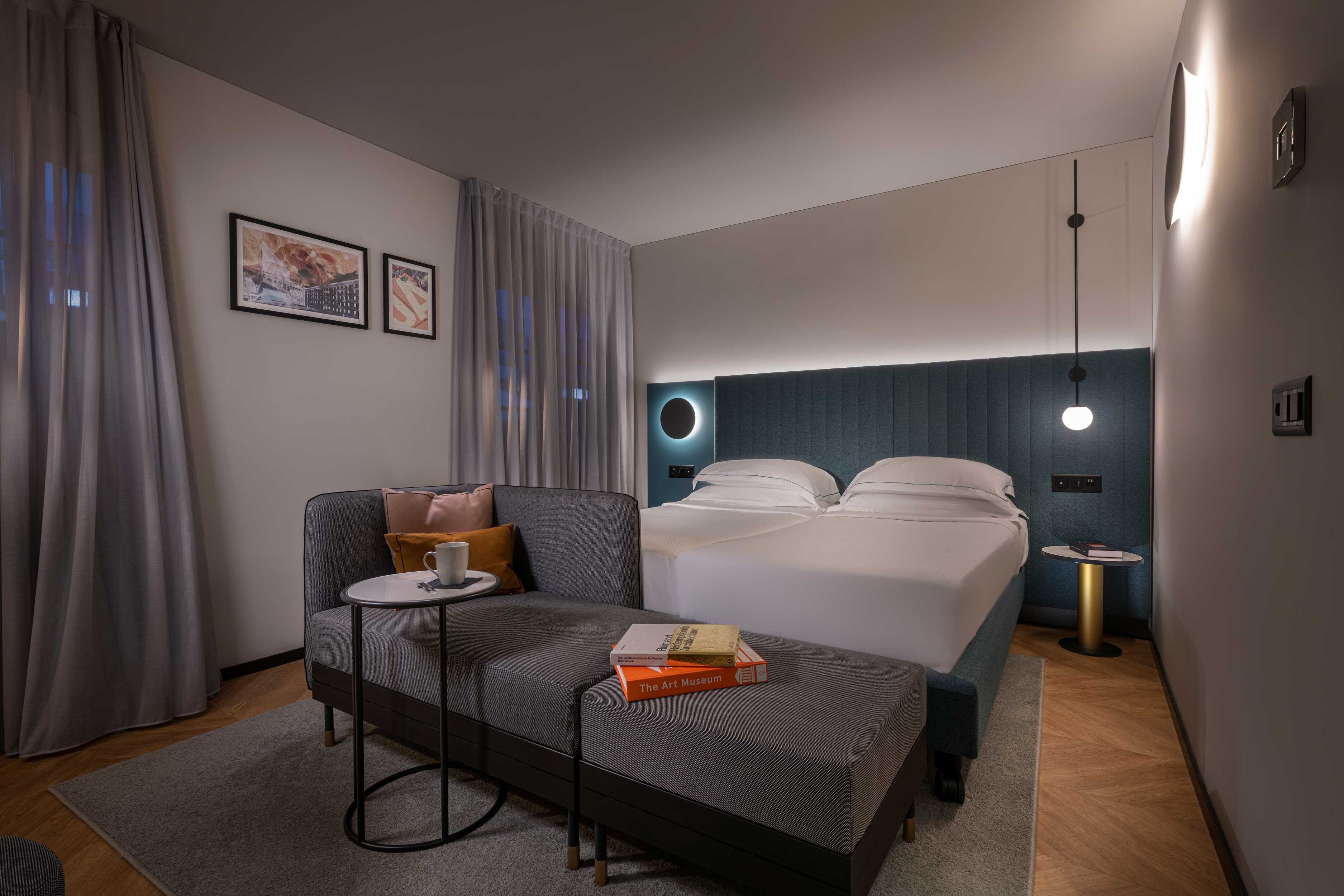 A modern hotel room with a bed, a sofa, a small table, and a bedside table. There are curtains, a wall-mounted light, and framed pictures on the wall.