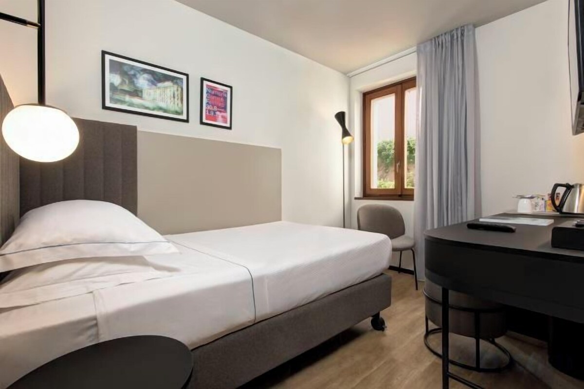 A hotel room with a bed, a desk, and a chair. There are two framed pictures on the wall, a window with curtains, and a lamp on the desk.