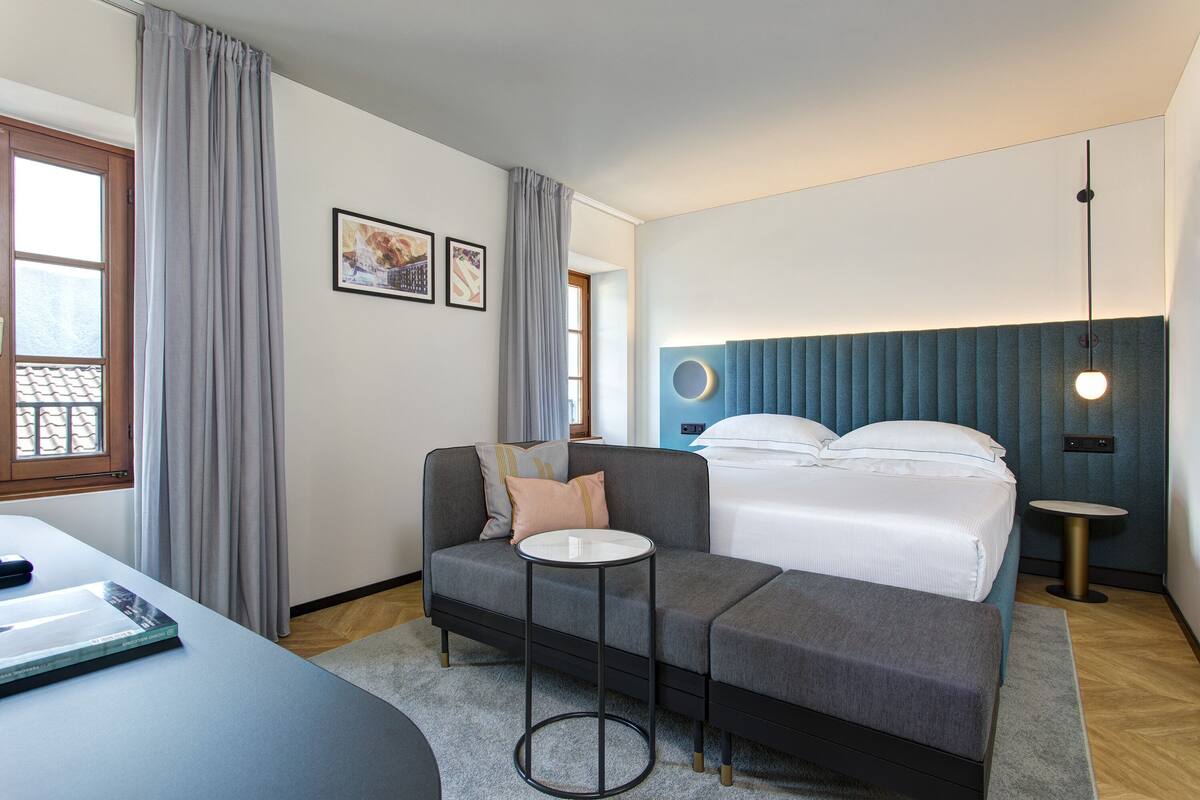 A modern hotel room with a sofa, a small round table, a bed with a headboard, and a bedside lamp.