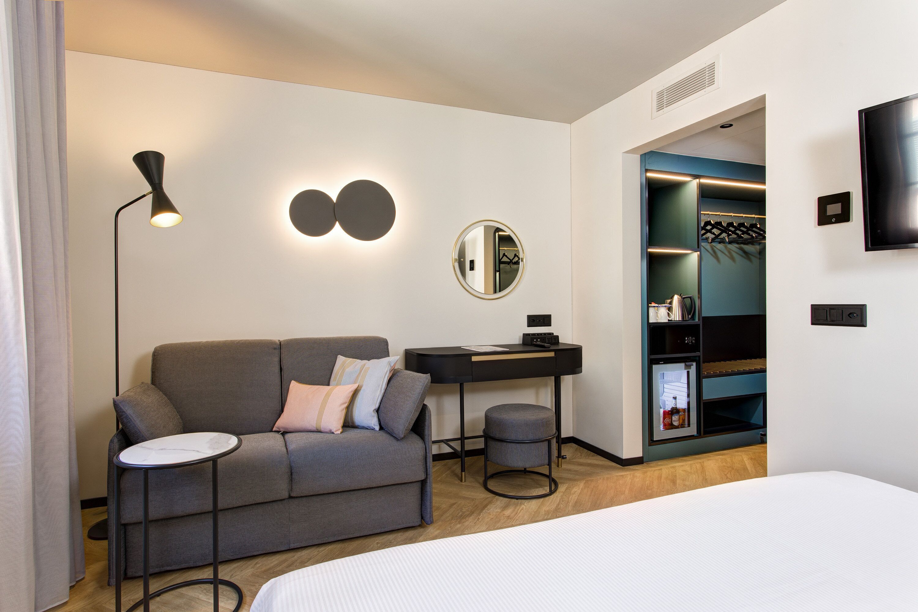Een moderne hotelkamer met een grijze bank, een klein rond tafeltje, een bureau, een spiegel, een kledingkast en een televisie.