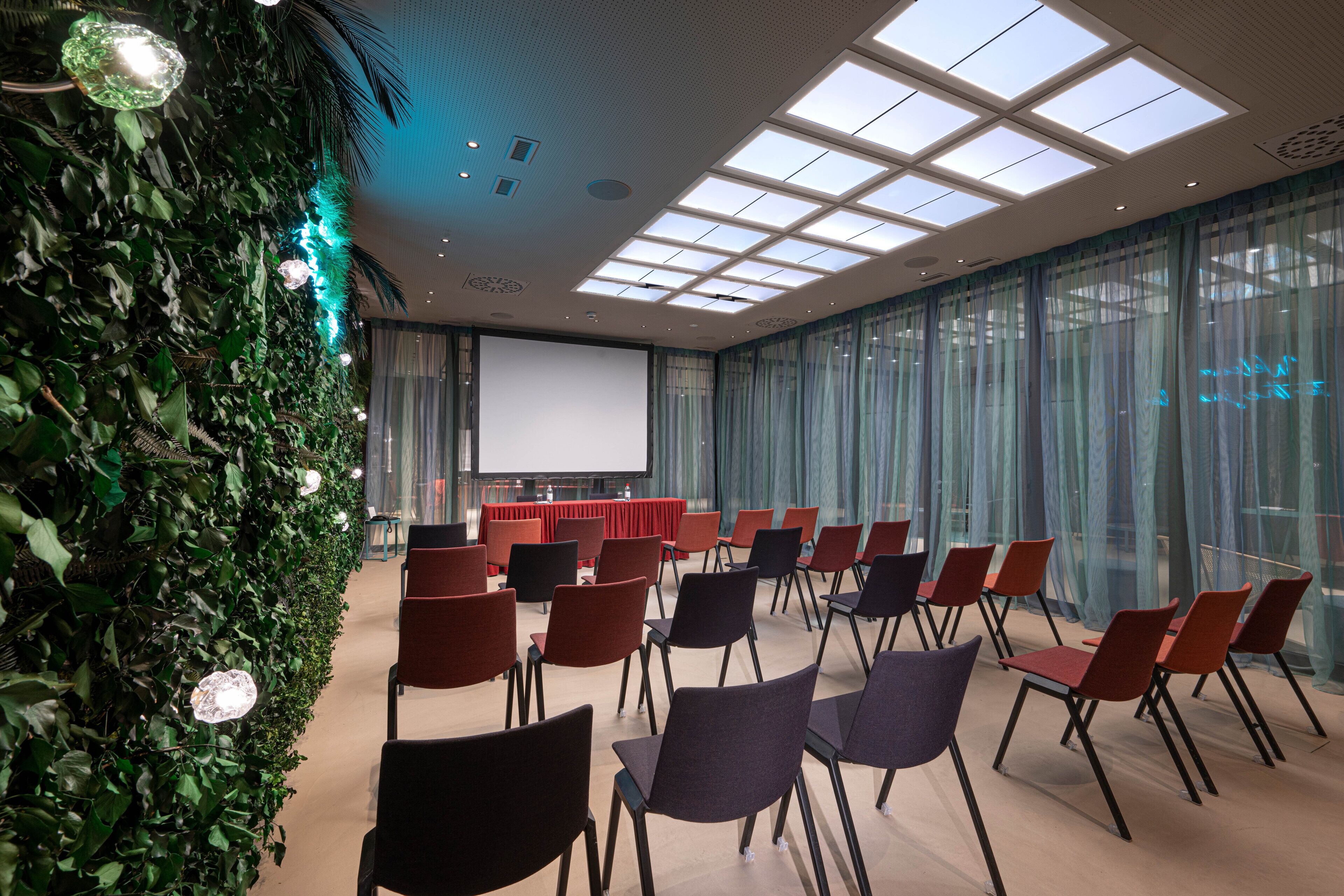 Une salle de conférence dotée d’un grand écran, de rangées de chaises et d’un mur vert orné de luminaires suspendus.