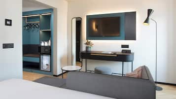 Et moderne hotelværelse med en seng, et skrivebord med et fladskærms-TV, en lille skammel, en gulvlampe og et walk-in closet med hylder og bøjlestang.