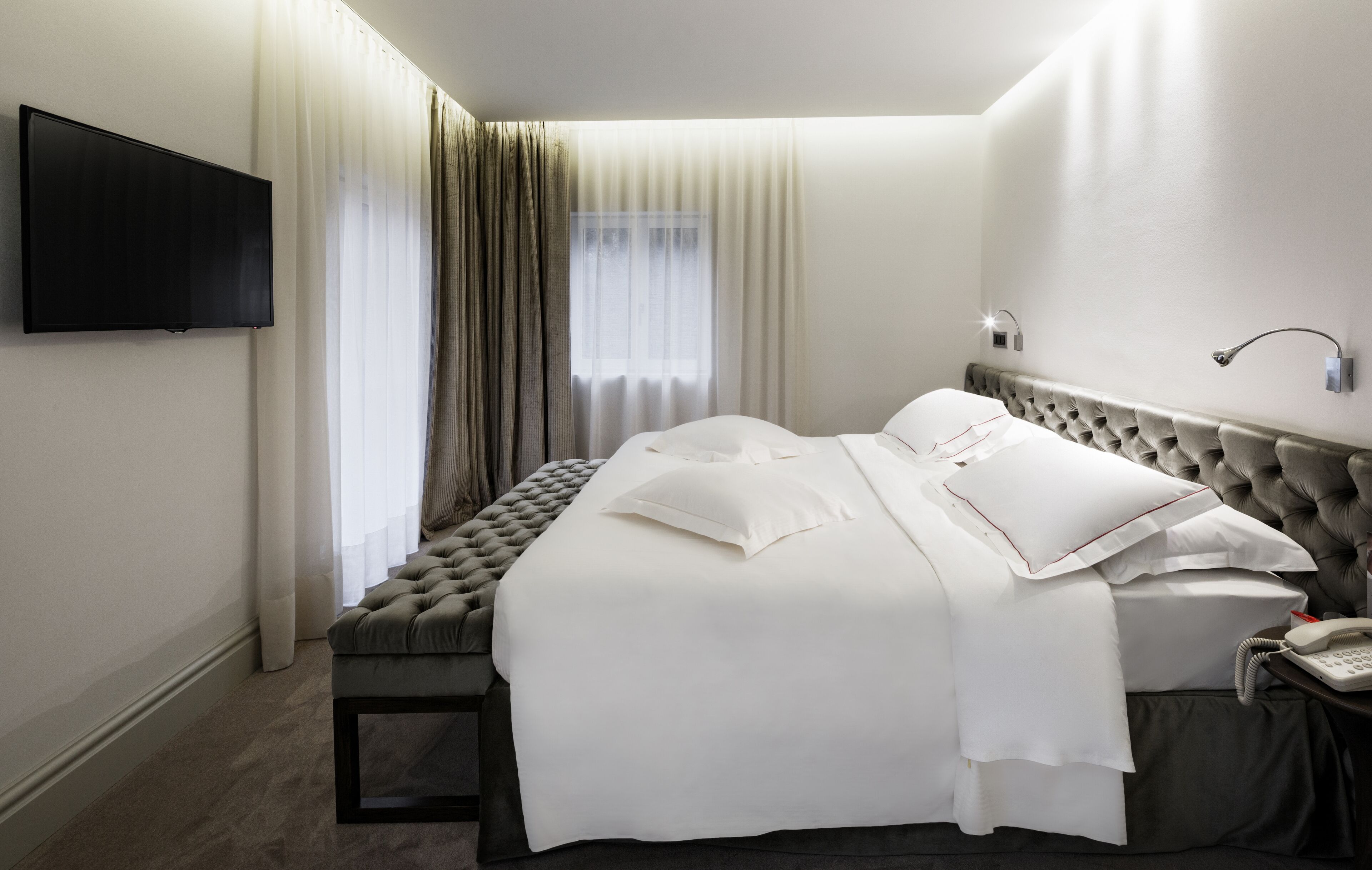 A hotel room with a large bed, a flat-screen TV mounted on the wall, a bedside table with a phone, and a window with sheer curtains.