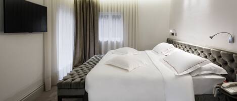 A hotel room with a large bed, a flat-screen TV mounted on the wall, a bedside table with a phone, and a window with sheer curtains.