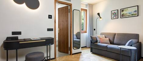 Una sala moderna con un sofá gris, una mesita redonda pequeña, un escritorio negro con una silla y un espejo.