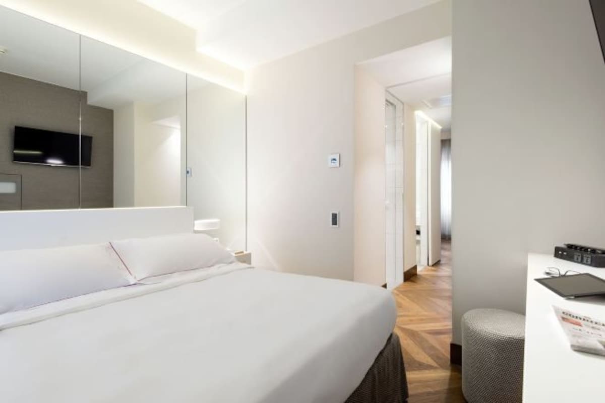 Una habitación de hotel con cama, escritorio, silla, televisor y espejo.