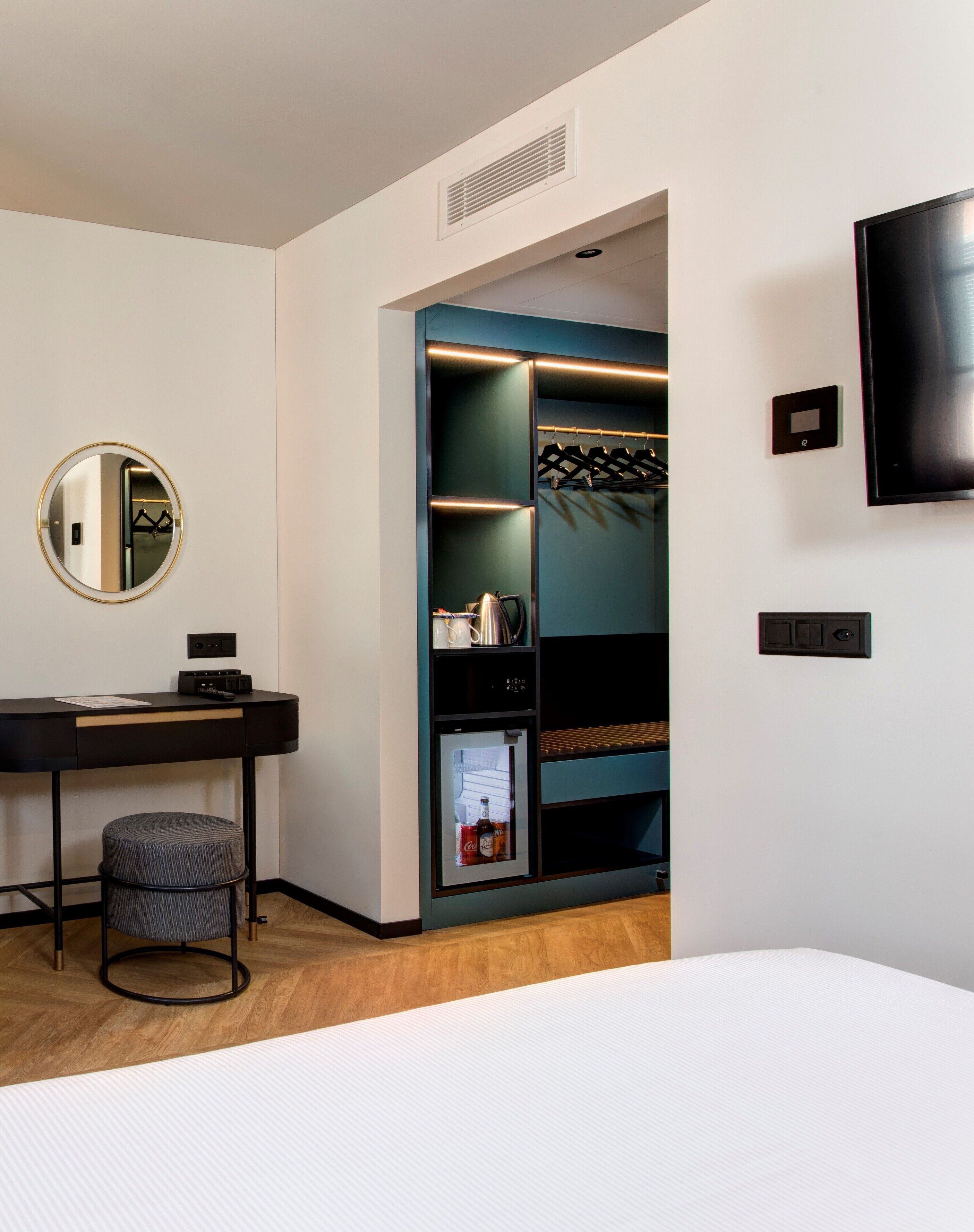 A modern hotel room with a bed, a desk with a chair, a minibar, a coat rack, and a TV mounted on the wall.