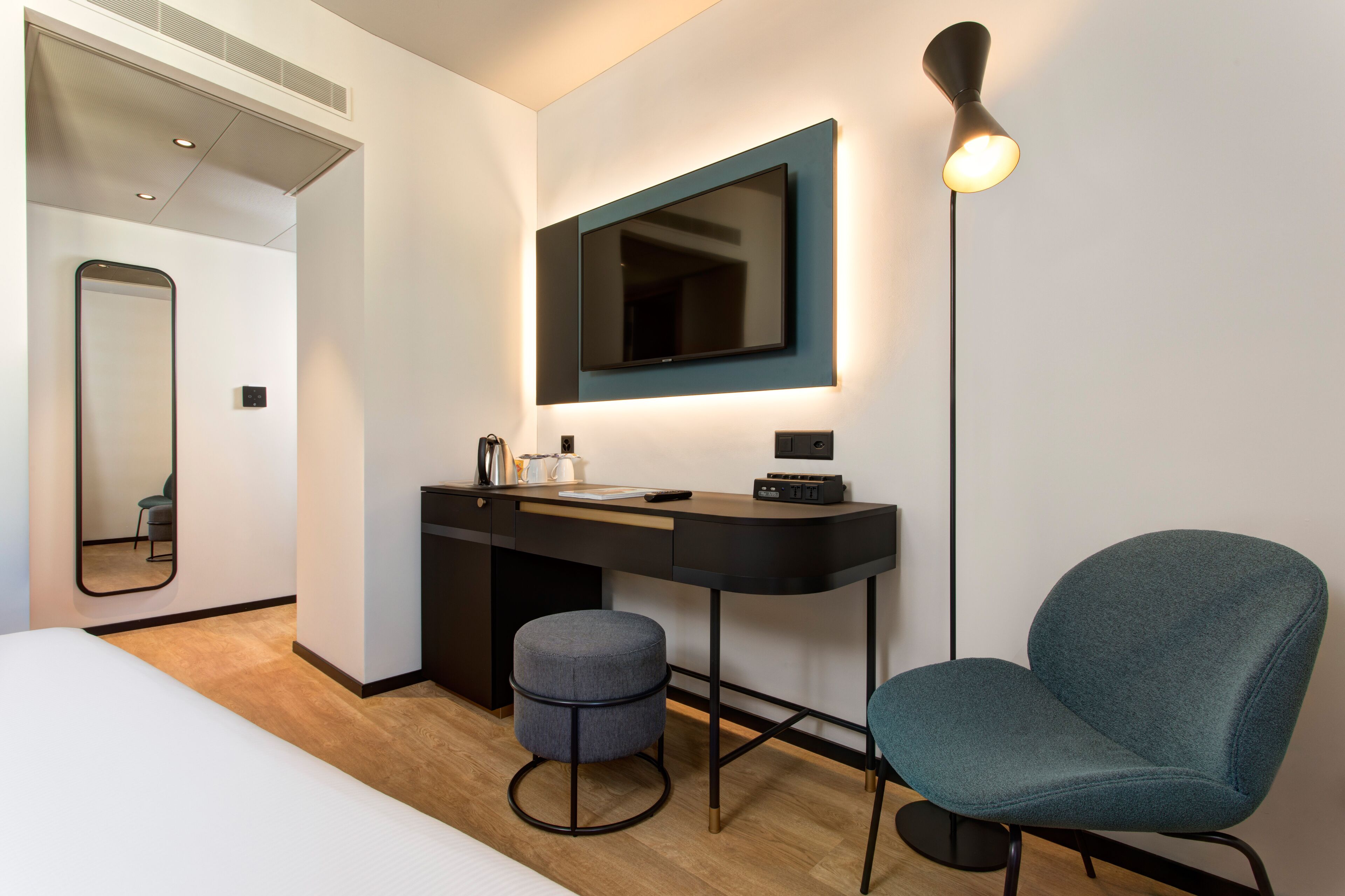 A modern hotel room with a bed, a desk with a mirror, a chair, and a flat-screen TV mounted on the wall.