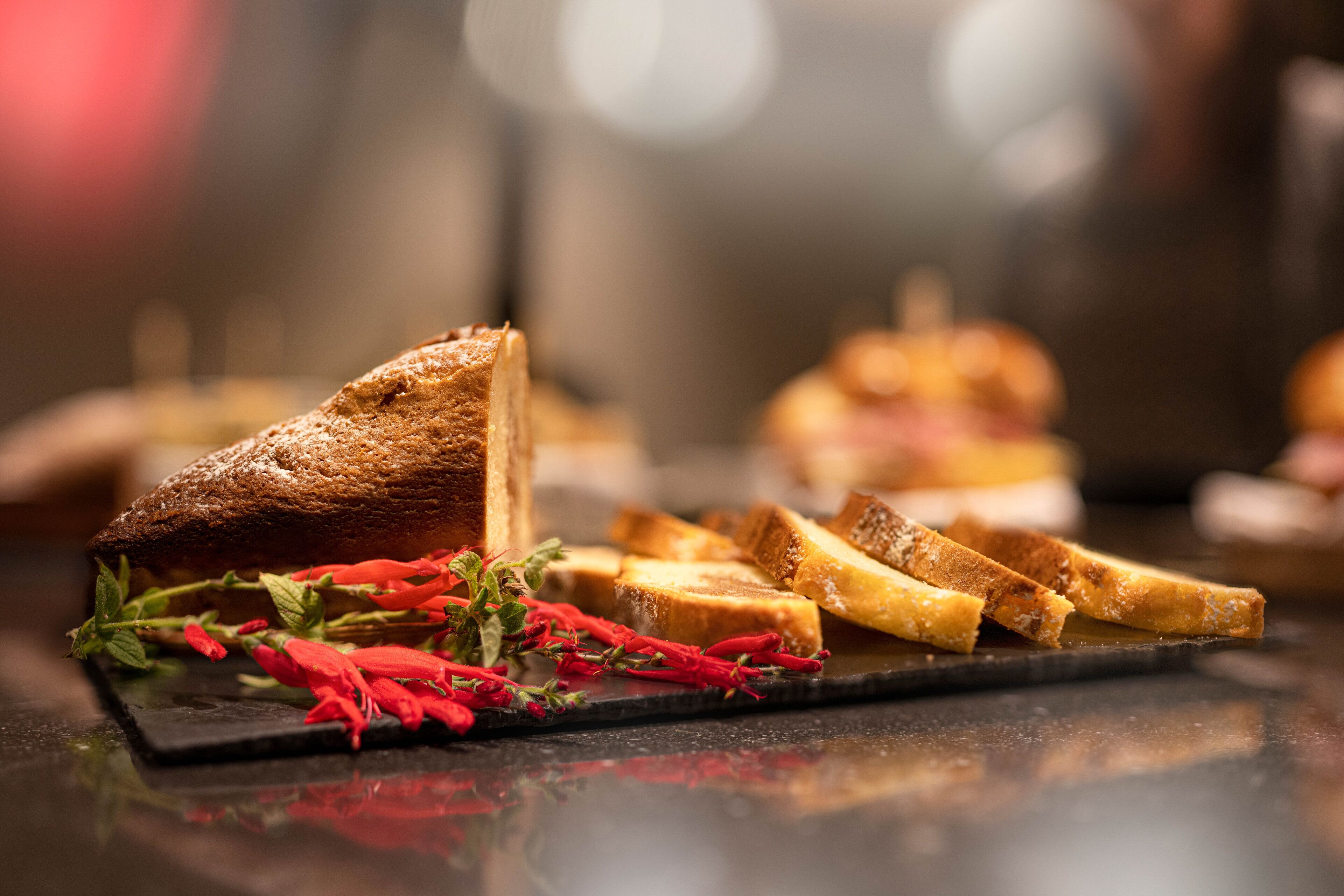 Eine Scheibe geröstetes Brot auf einer dunklen Schieferplatte, mit roten Blumen und einem ganzen Brot im Hintergrund.